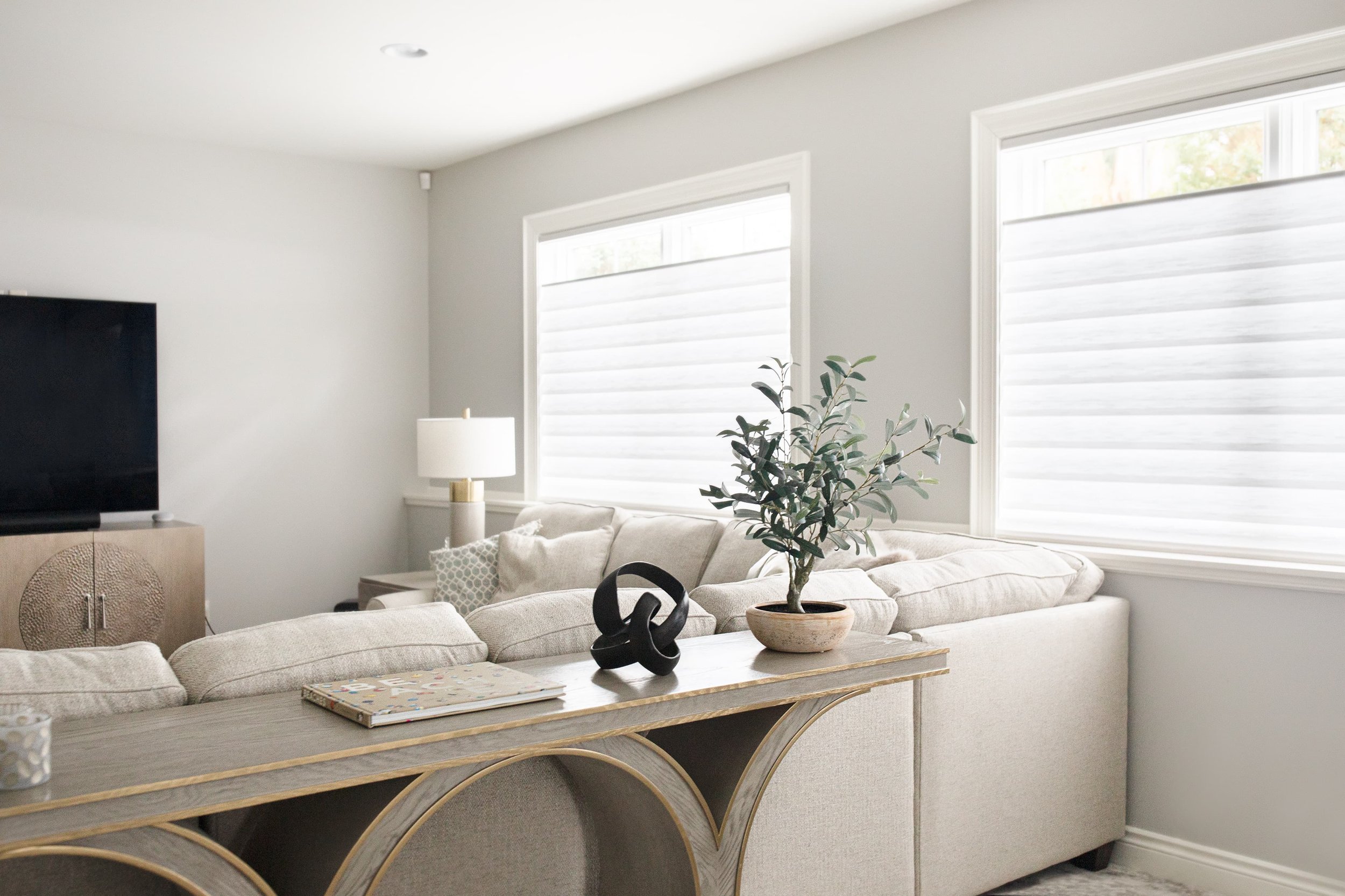 Neutral living room with light fabric sofa, wood console table, decorative objects, potted plant, and white light-filtering window shades in a calm, modern space.