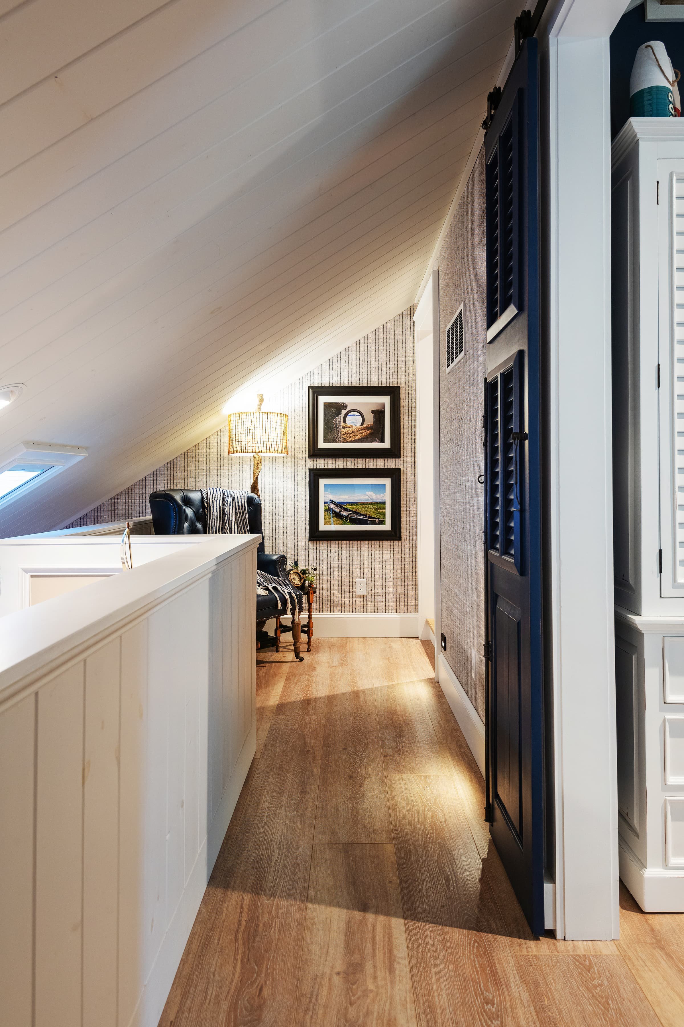 Upper-level hallway with sloped ceiling, wood floors, blue door, and a reading nook with a leather chair, floor lamp, and framed artwork.