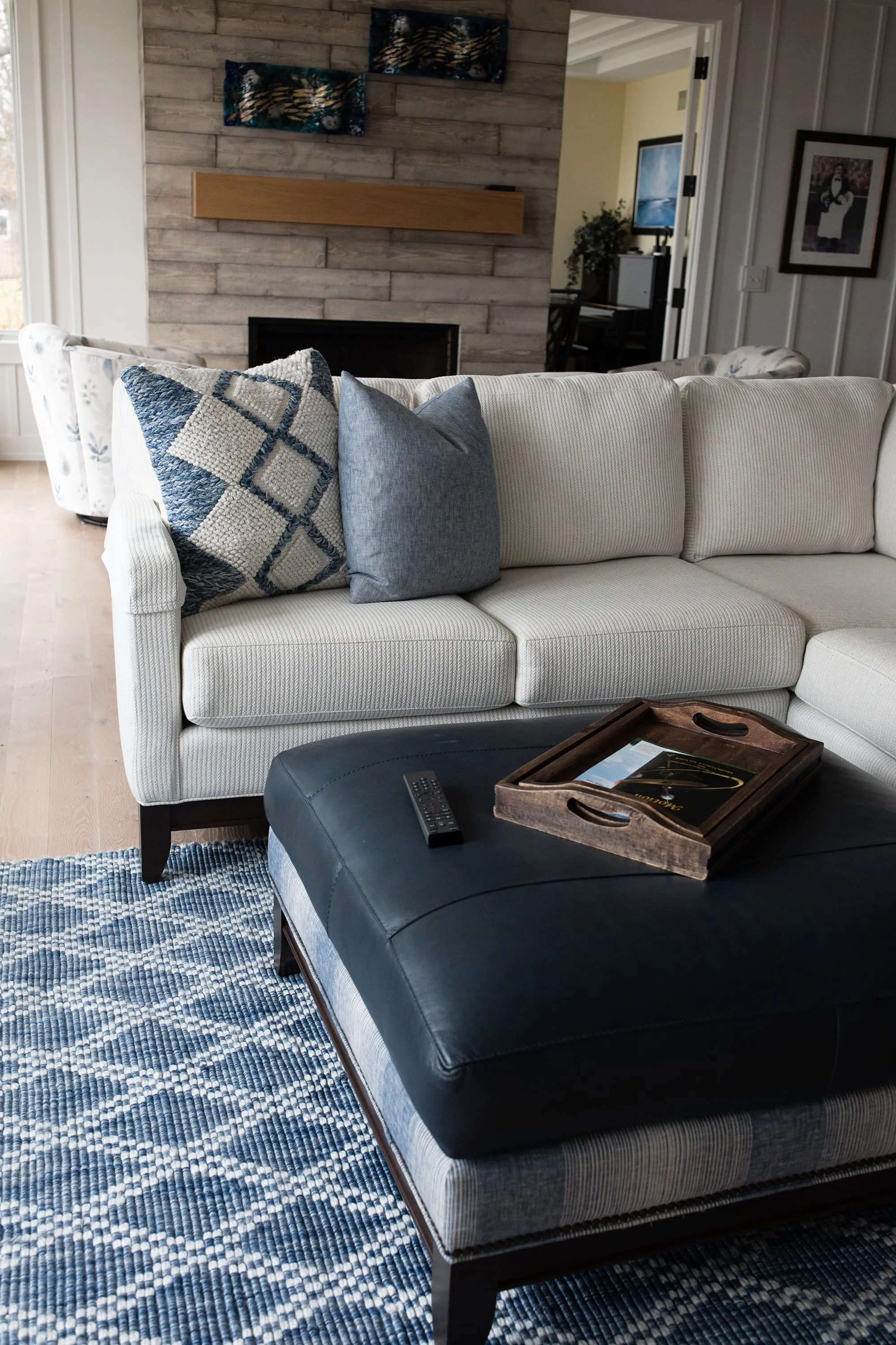 Neutral sectional sofa with blue accent pillows, dark leather ottoman, wooden tray, and blue patterned area rug in a bright, modern living room.