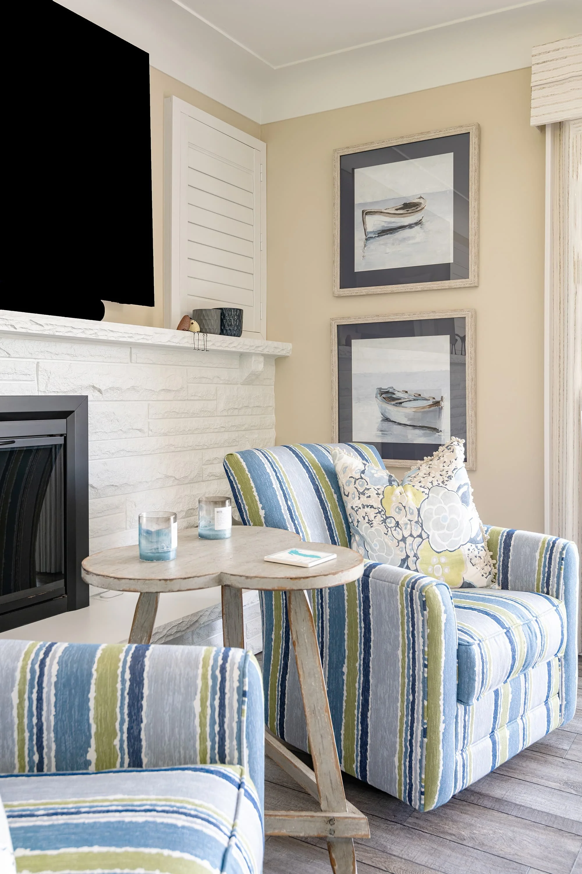 Living area with blue and green striped armchairs, small wood side table, white brick fireplace, and framed boat artwork in a bright, relaxed setting.
