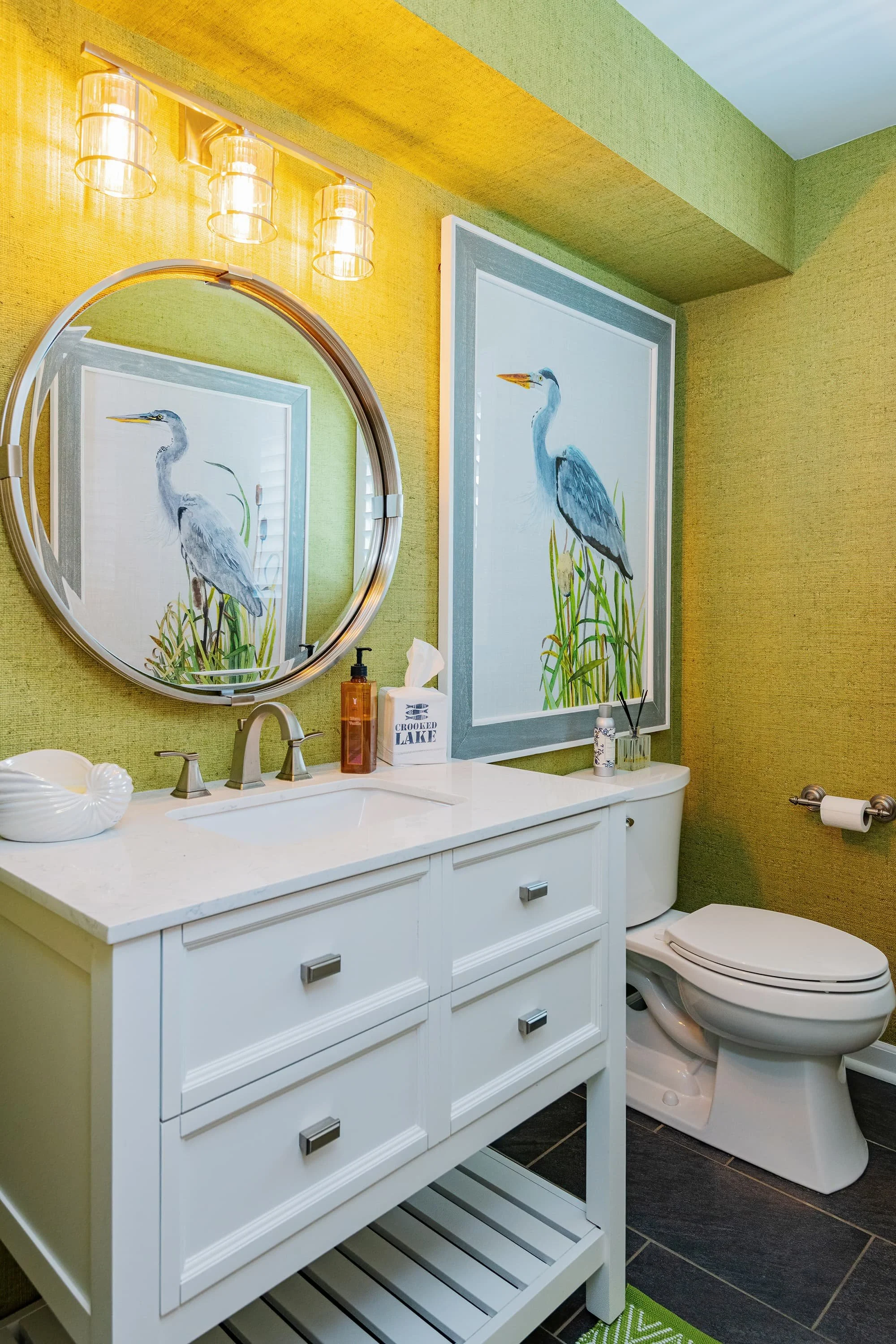 Powder bathroom with green grasscloth walls, white vanity, round mirror, brass lighting, and framed bird artwork in a bold, colorful space.