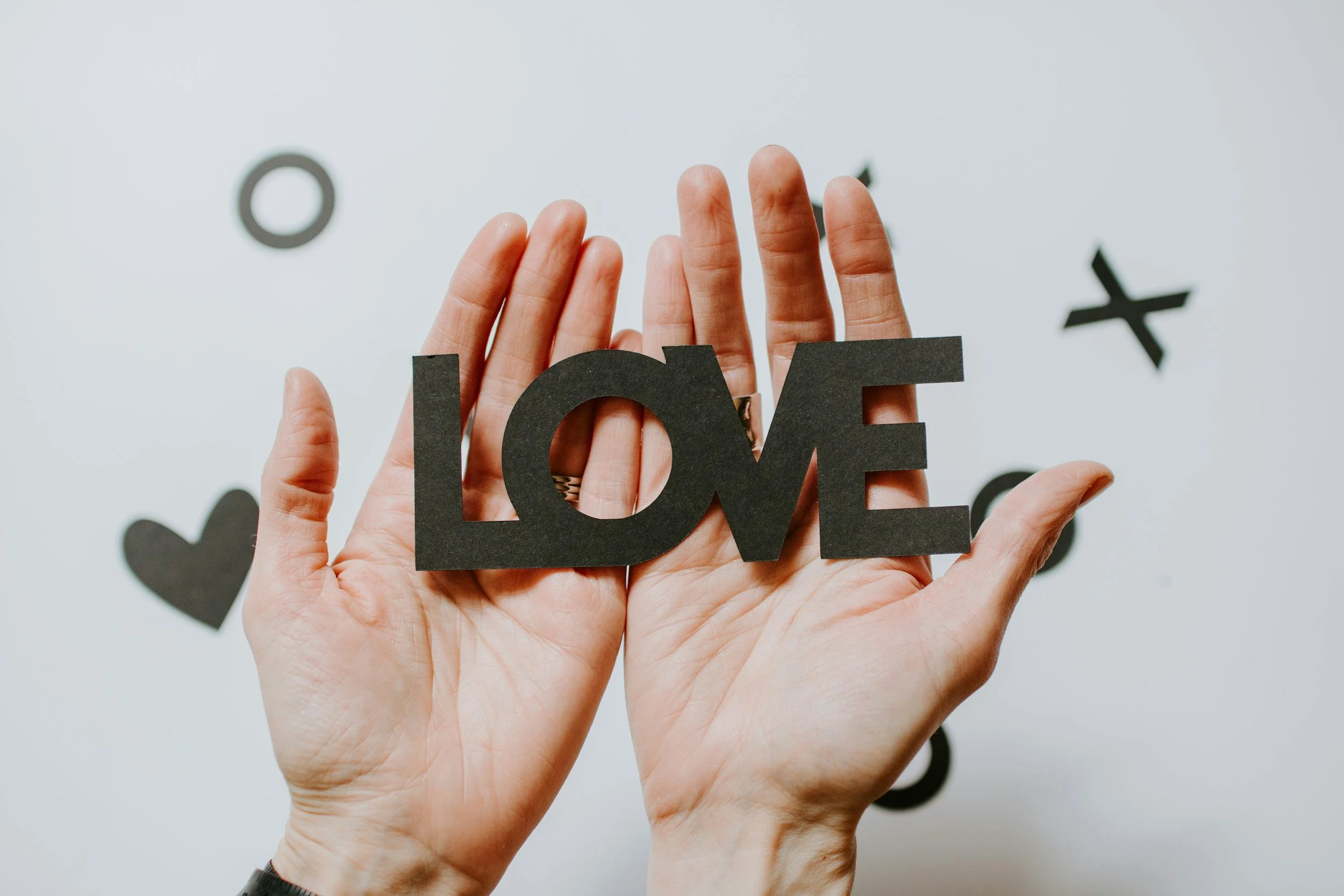Two hands holding a black cut-out of the word 'LOVE' in front of a white wall with black paper cut-outs of hearts, circles, and X shapes.