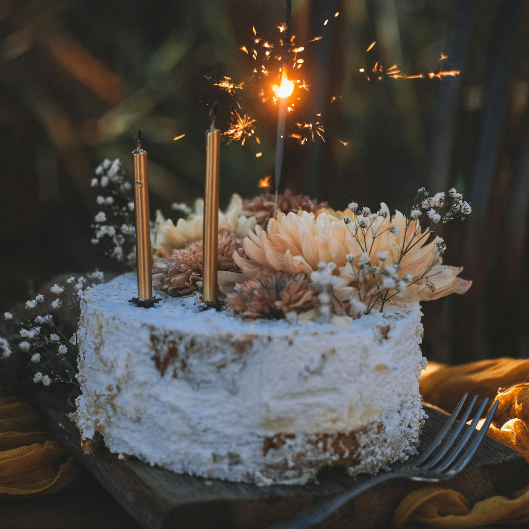 A cake decorated with flowers and sparklers, with two lit yellow candles, on a wooden surface with a fork nearby.