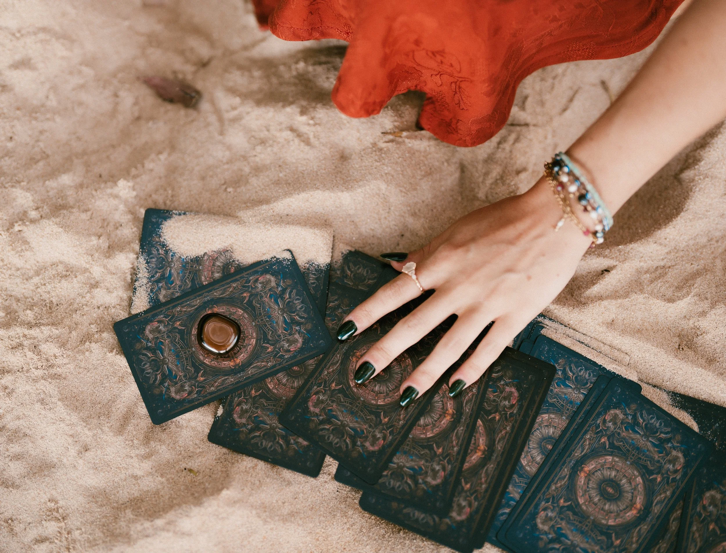 A person with painted black nails and multiple colorful bracelets is placing or reaching for tarot or oracle cards on sand, with a cloth partially visible underneath.