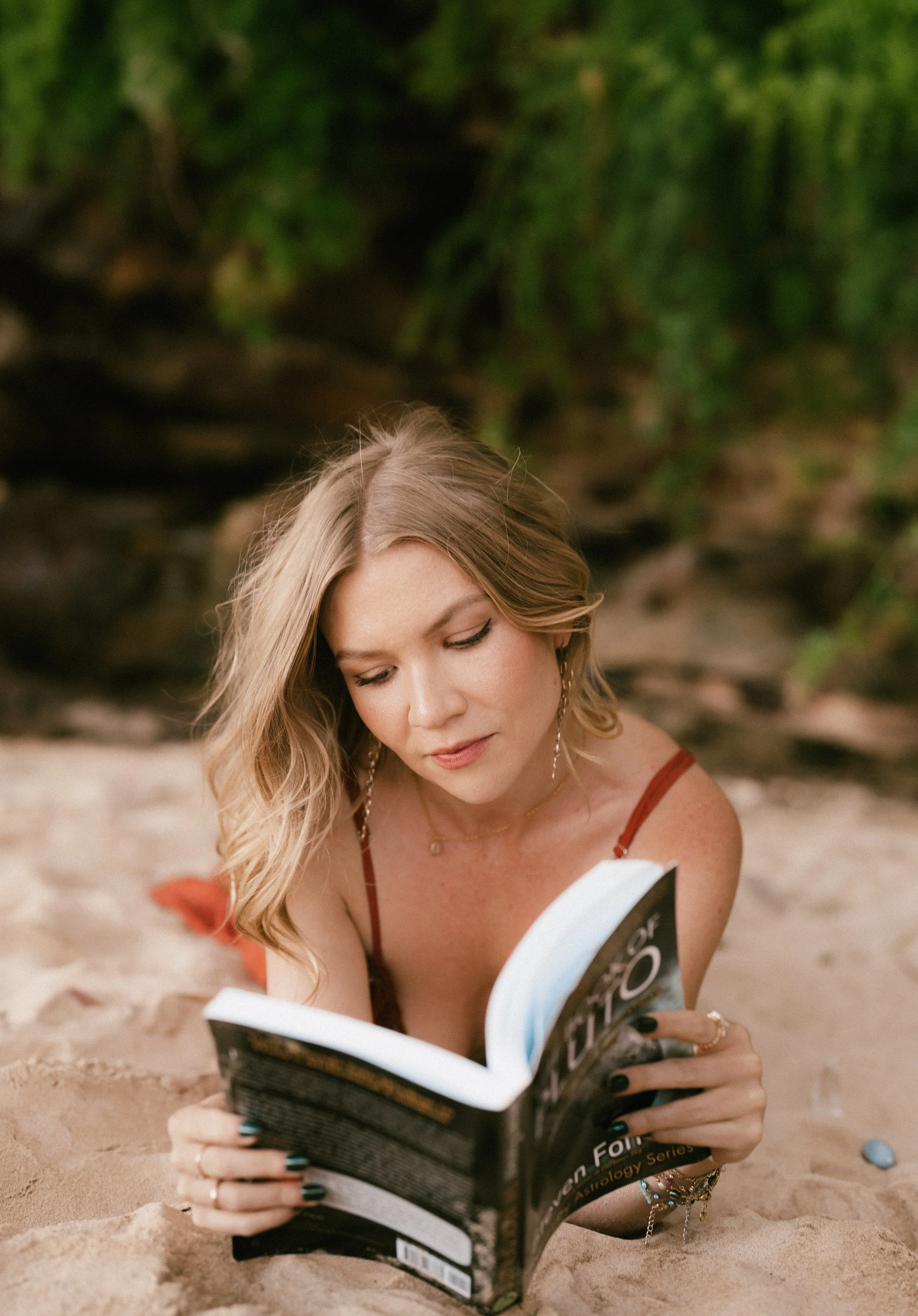 A woman with wavy blonde hair reading an astrology book while lying on sandy ground outdoors, with green foliage and rocks in the background.