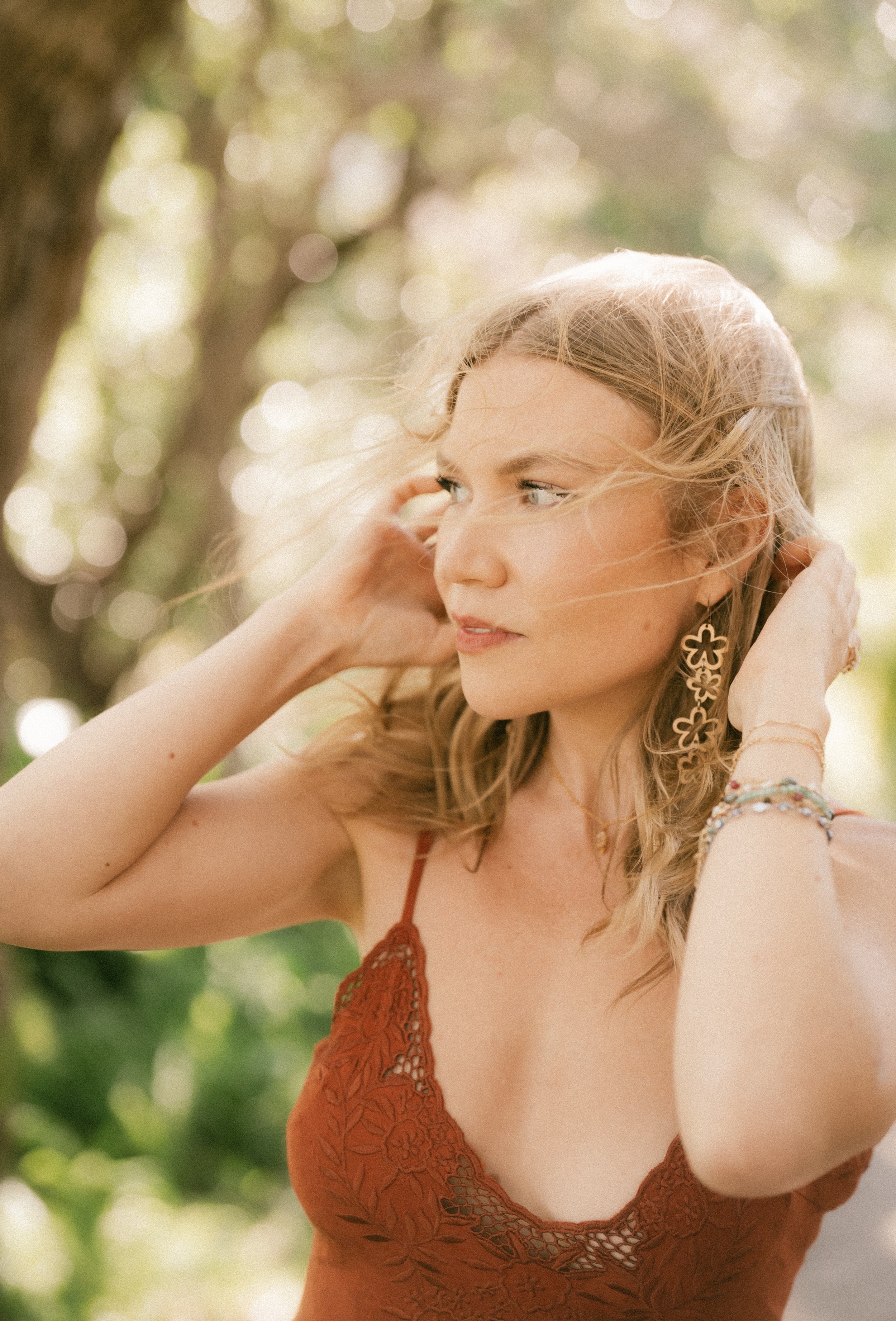 A woman with blonde wavy hair wearing floral earrings, multiple bracelets, and an orange lace dress, standing outdoors with sunlight filtering through trees, looking to the side.