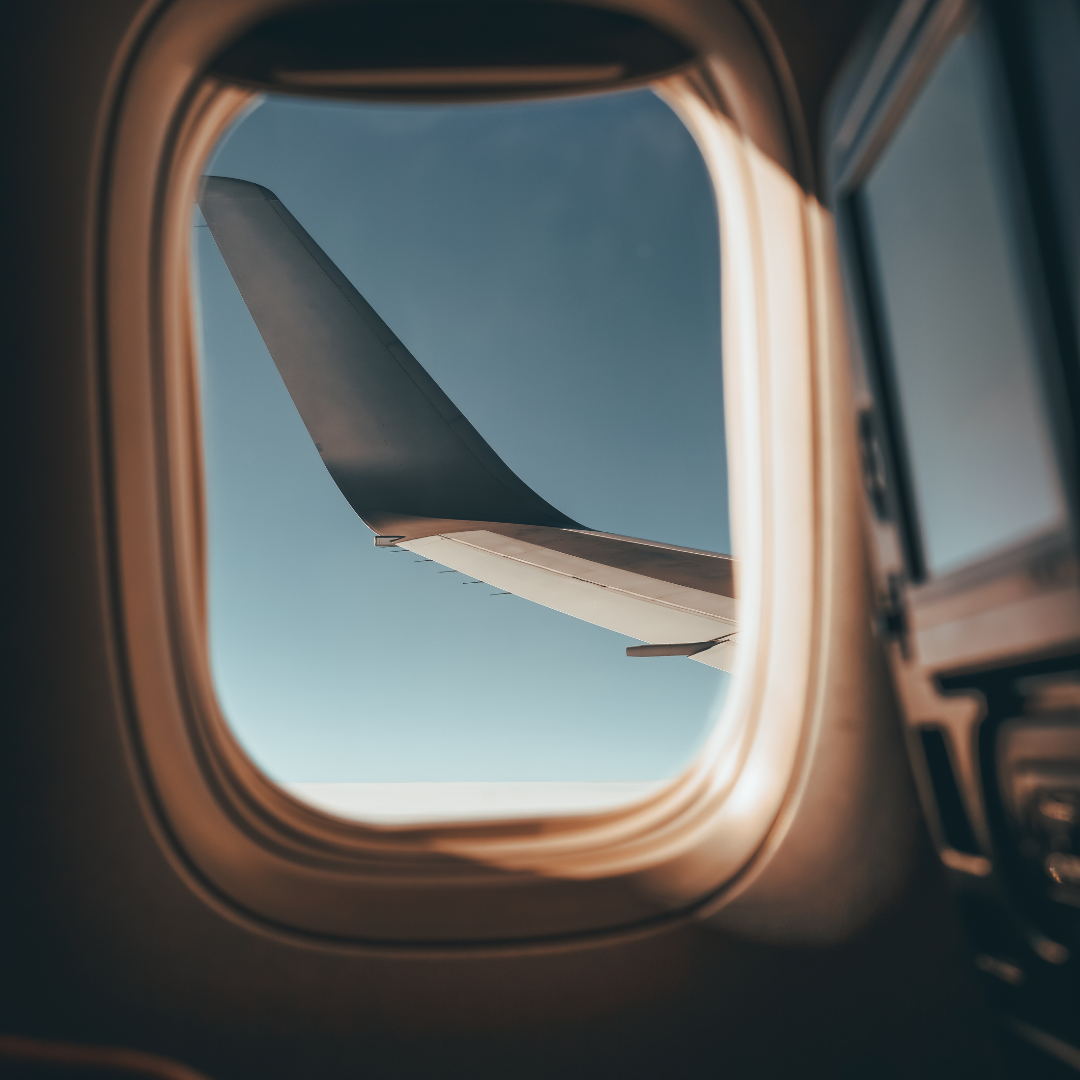View of an airplane wing through an aircraft window during flight, with blue sky visible outside.