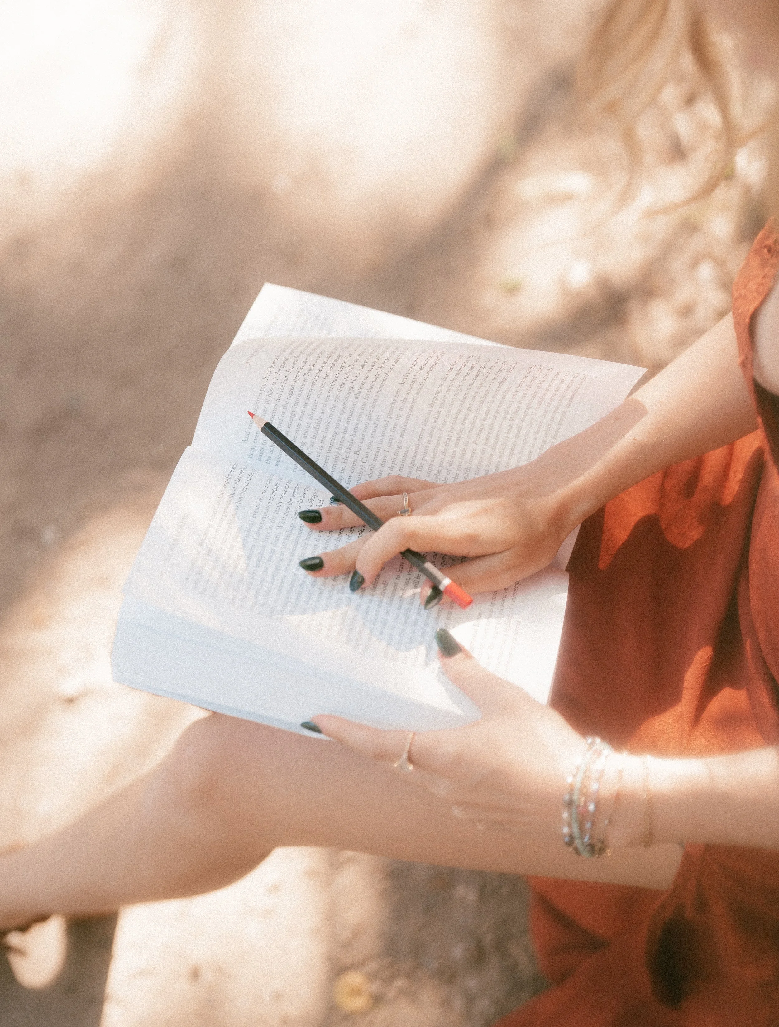 A person holding an astrology open book and a pen, sitting outdoors with sunlight and dirt ground visible.