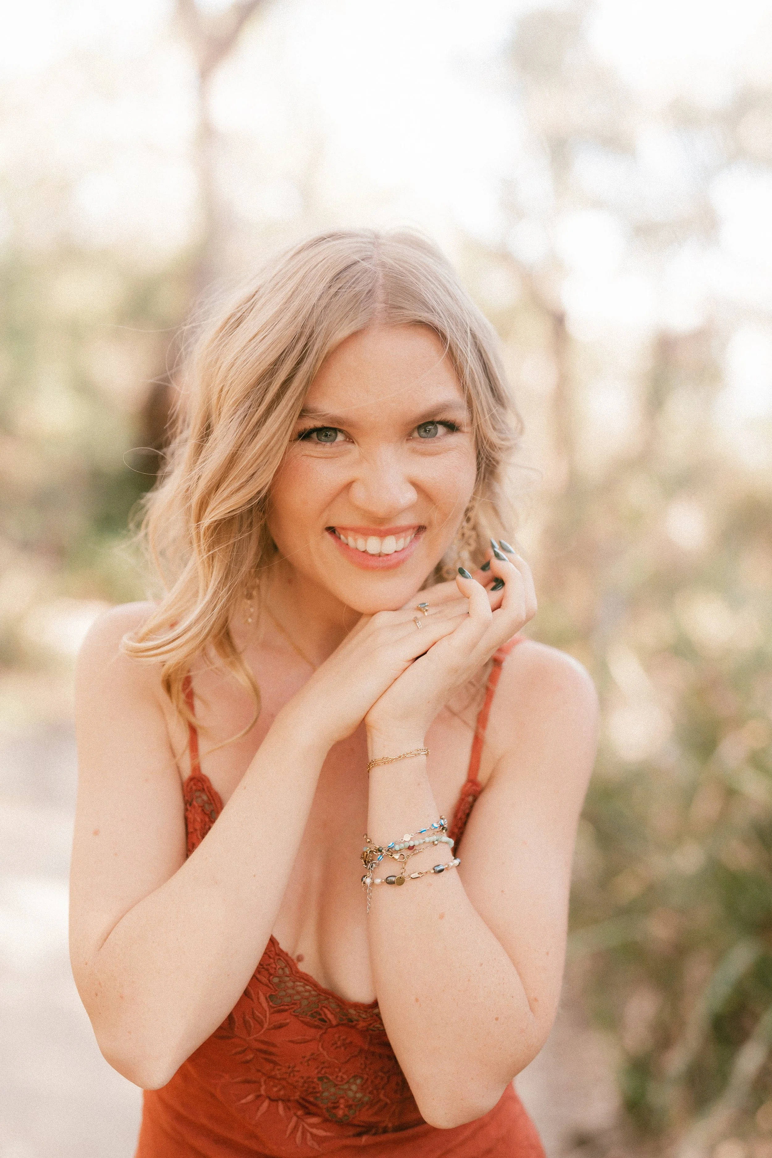 A woman with blonde hair in loose waves, smiling and posing outdoors, wearing a rust-colored dress with lace details and multiple bracelets on her wrist.