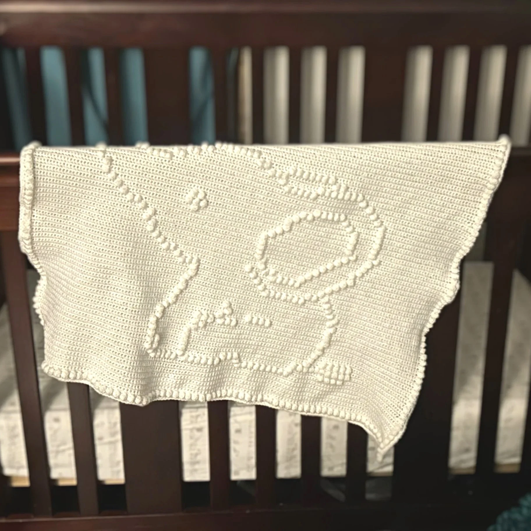 White crochet blanket with bobble stitch design of a baby elephant hanging on a dark wooden crib railing.
