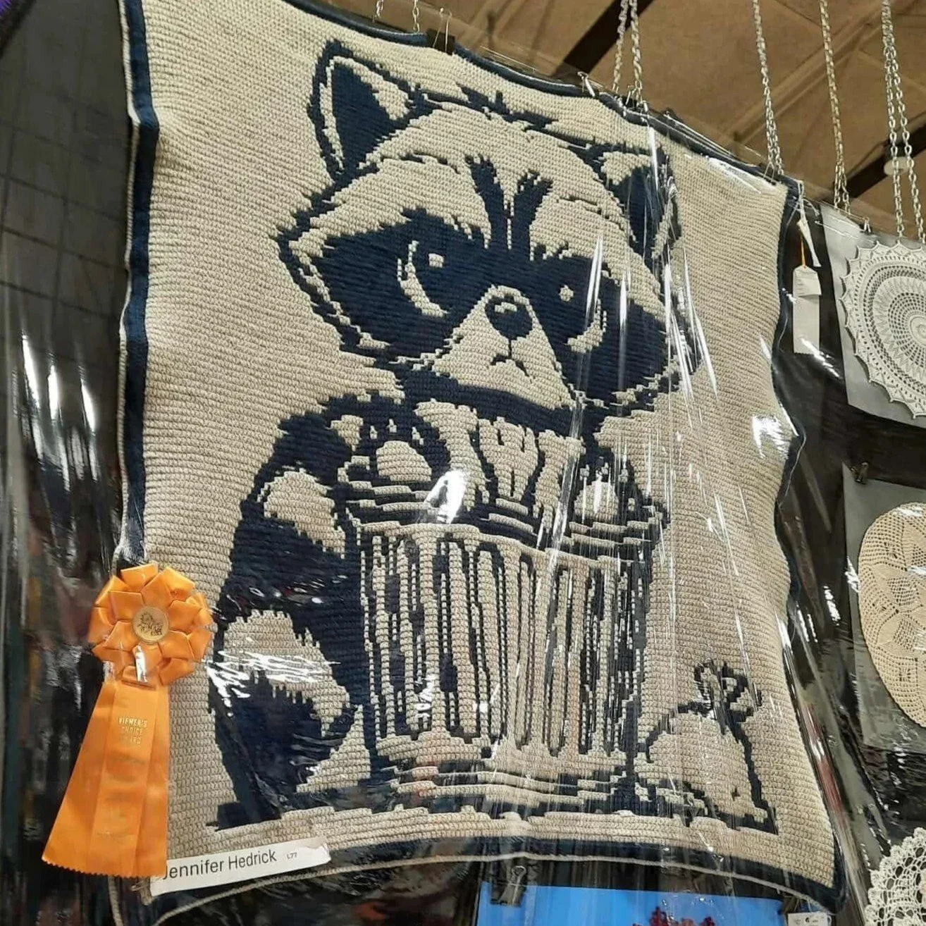 Reversible Mosaic style crocheted blanket featuring artwork of a raccoon sitting inside a trash can displayed with a ribbon on a wall at the Maryland State Fair.