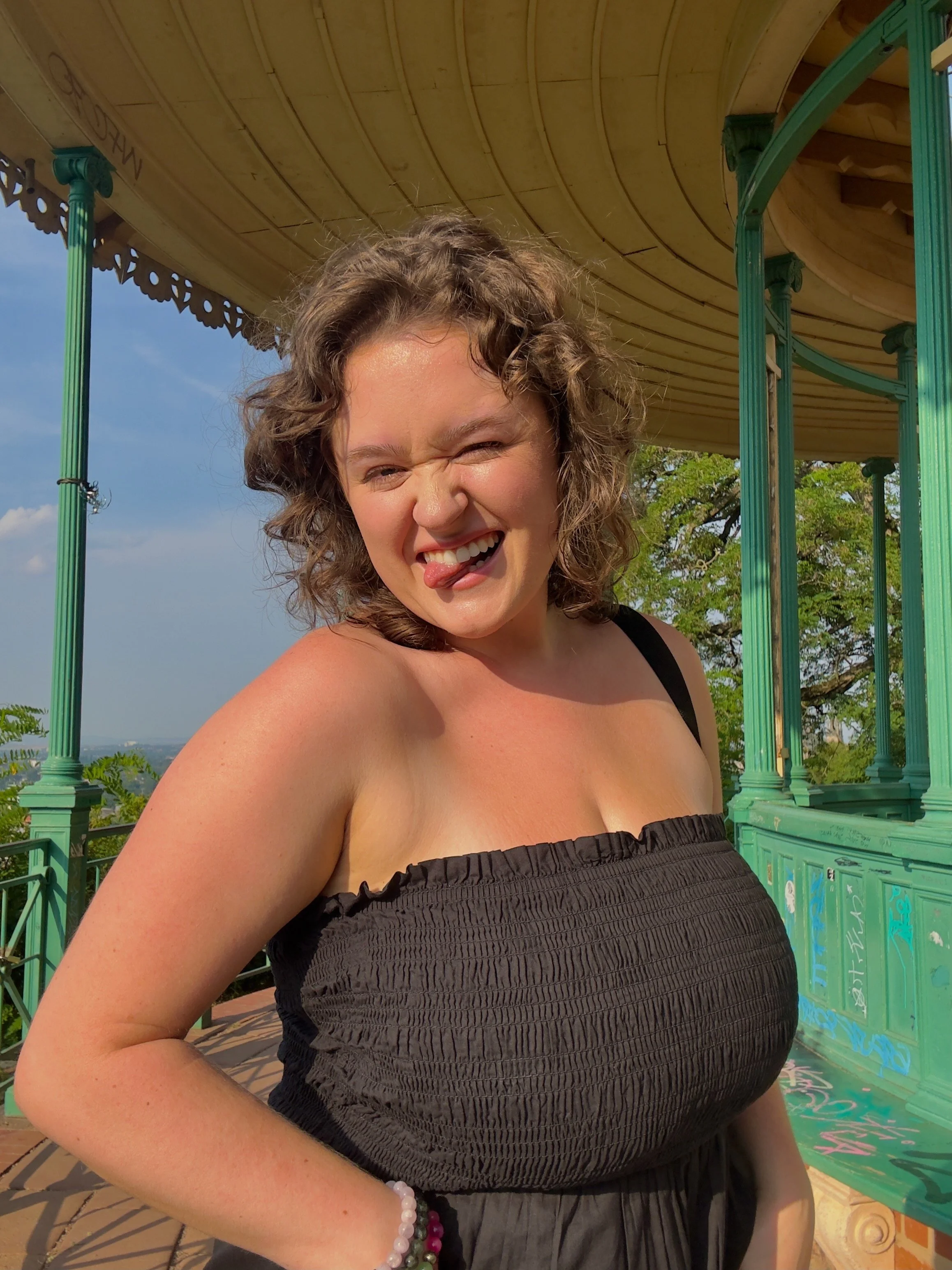 A woman with curly hair making a playful face with her tongue out and eyes squinted, standing outdoors near a green gazebo under a blue sky.