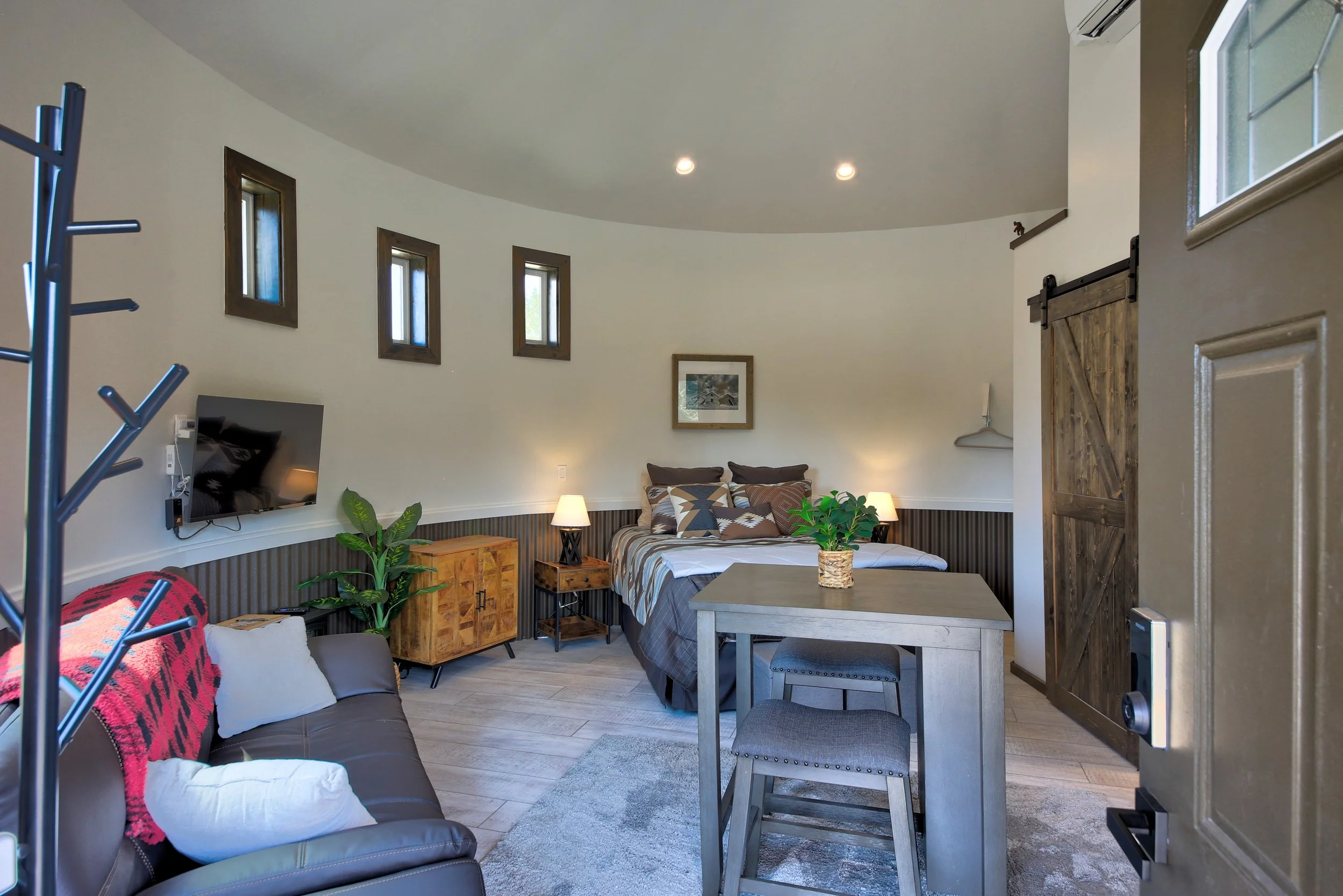 Cozy bedroom with a bed, side tables with lamps, a small table with a plant, a sofa with cushions and blanket, a wall-mounted TV, and a rustic sliding barn door in a modern, well-lit room.