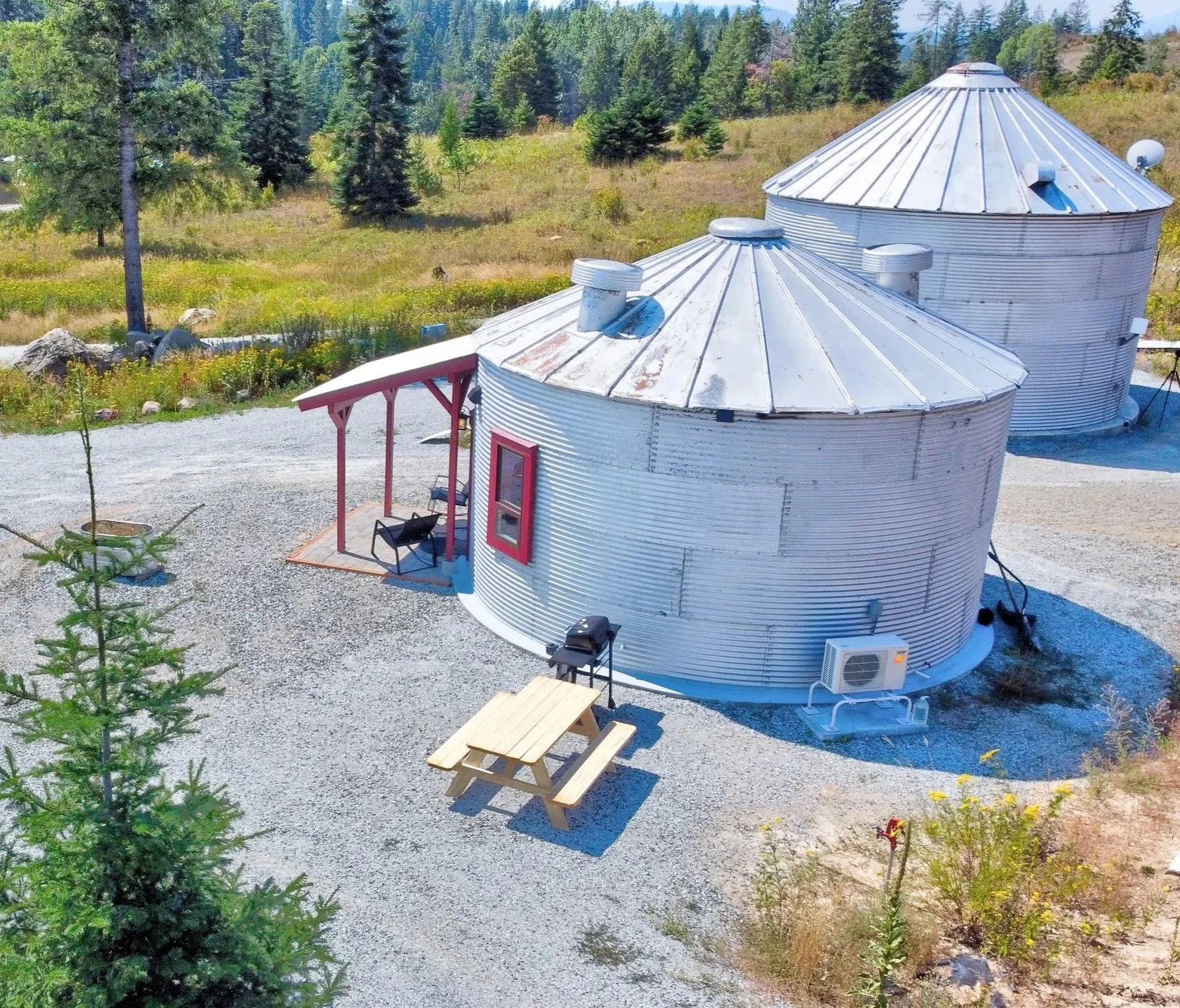 Two round metal silo structures with conical roofs on a gravel yard, surrounded by trees and hillside in the background.