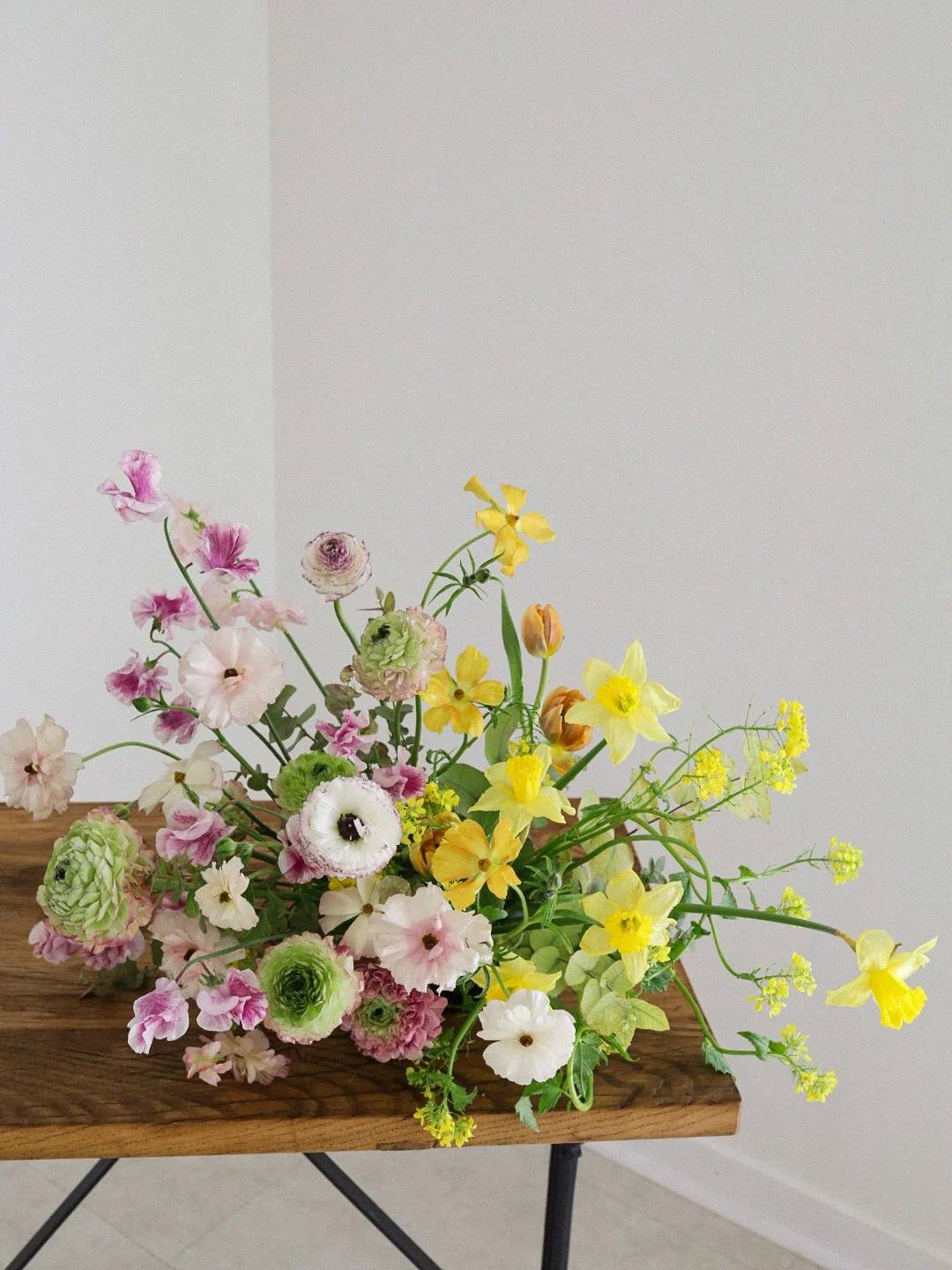 Colorful assortment of fresh flowers in a rustic wooden vase on a dark metal table against a plain light-colored wall.