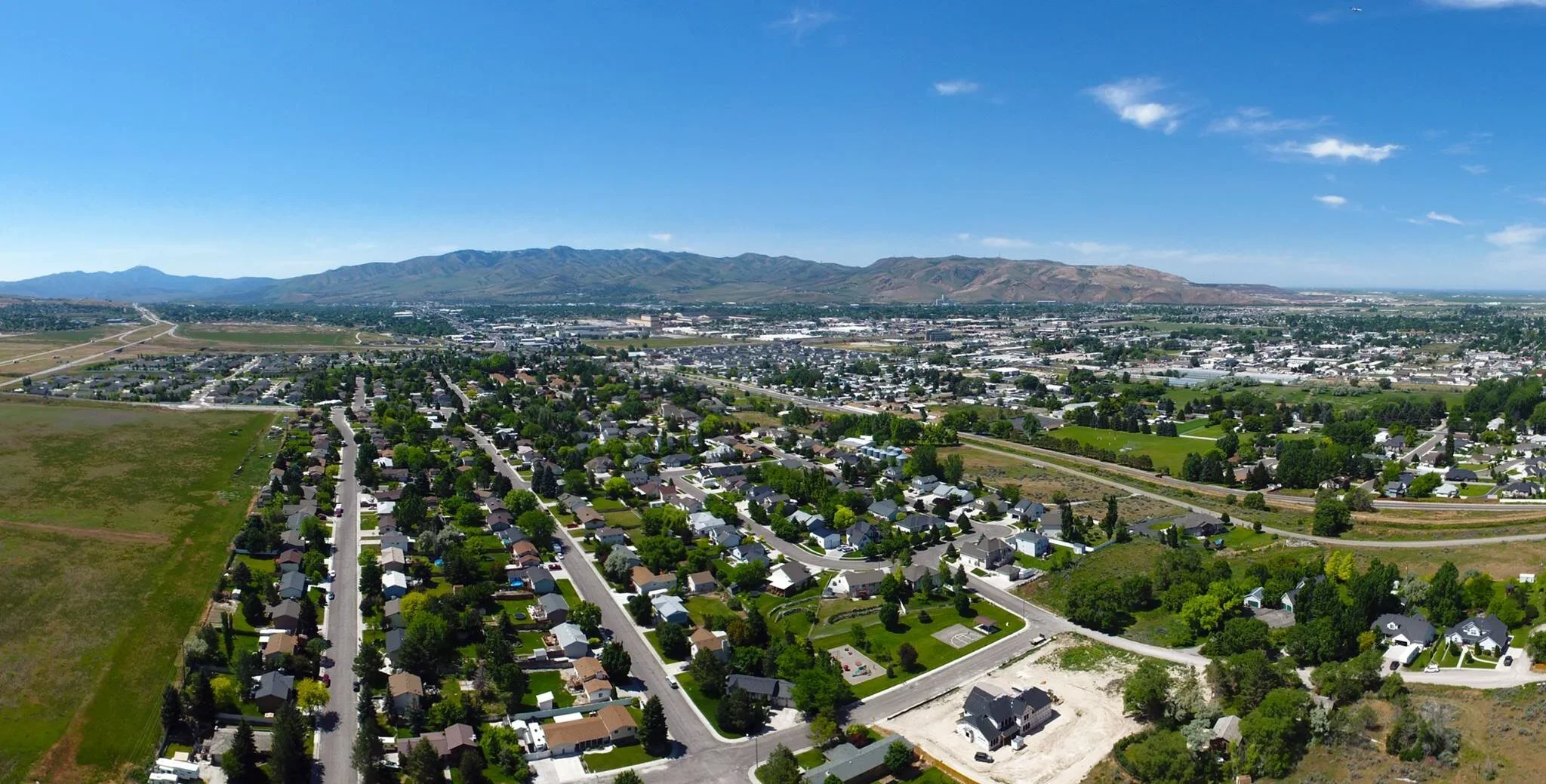 Aerial view of a suburban neighborhood with houses and trees, landscape includes mountains in the background, under a clear blue sky.