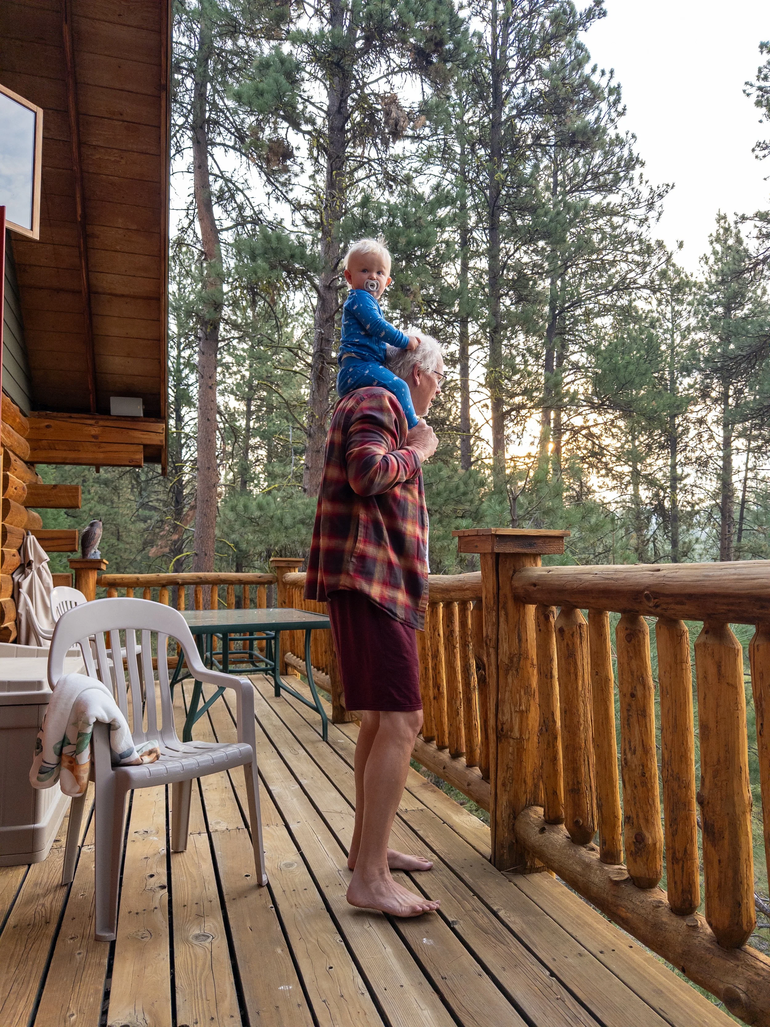 An older man with white hair carrying a young child on his shoulders on a wooden deck outside, surrounded by tall pine trees at sunset.