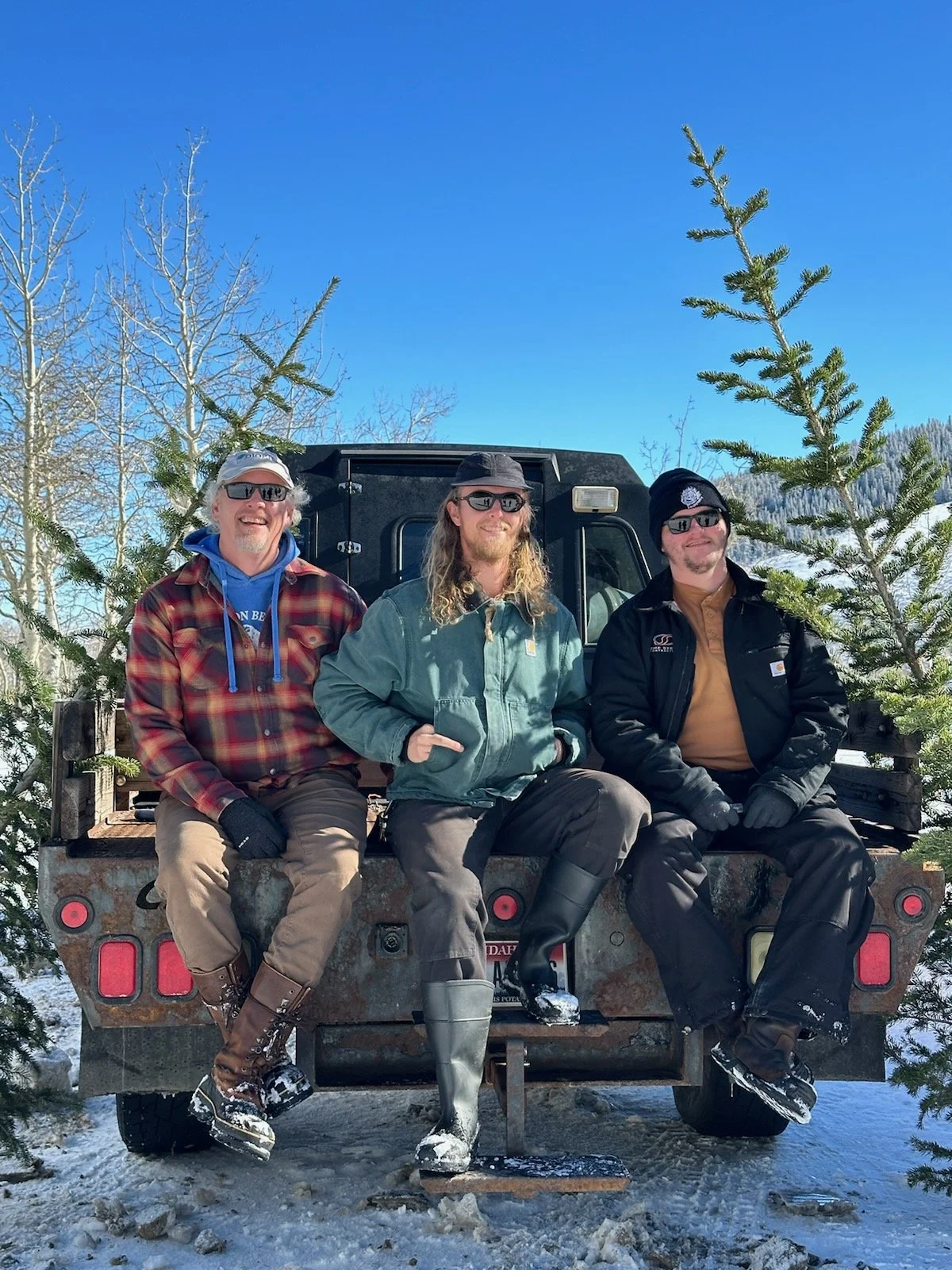 Three men sitting on the back of a flatbed truck in a snowy outdoor setting with small pine trees and a clear blue sky, wearing winter clothing and sunglasses.