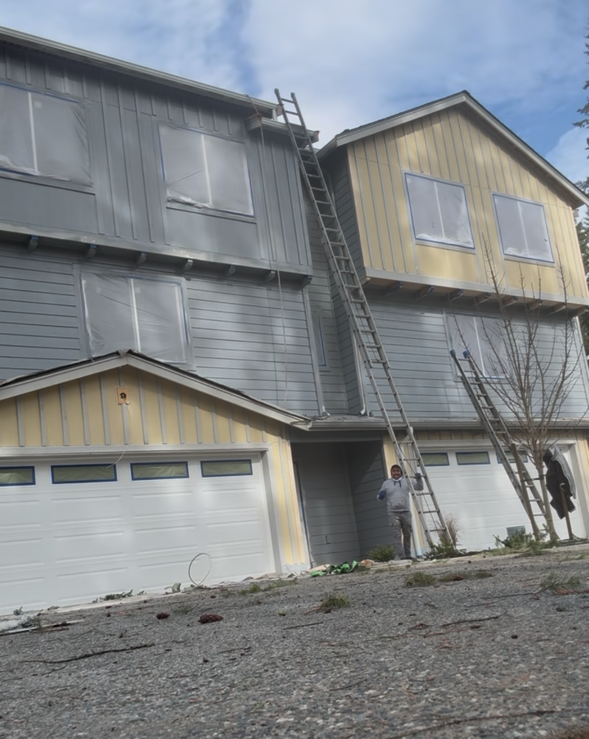 A two-story house with blue and yellow siding being painted, with ladders leaning against the building and a person standing near the entrance.