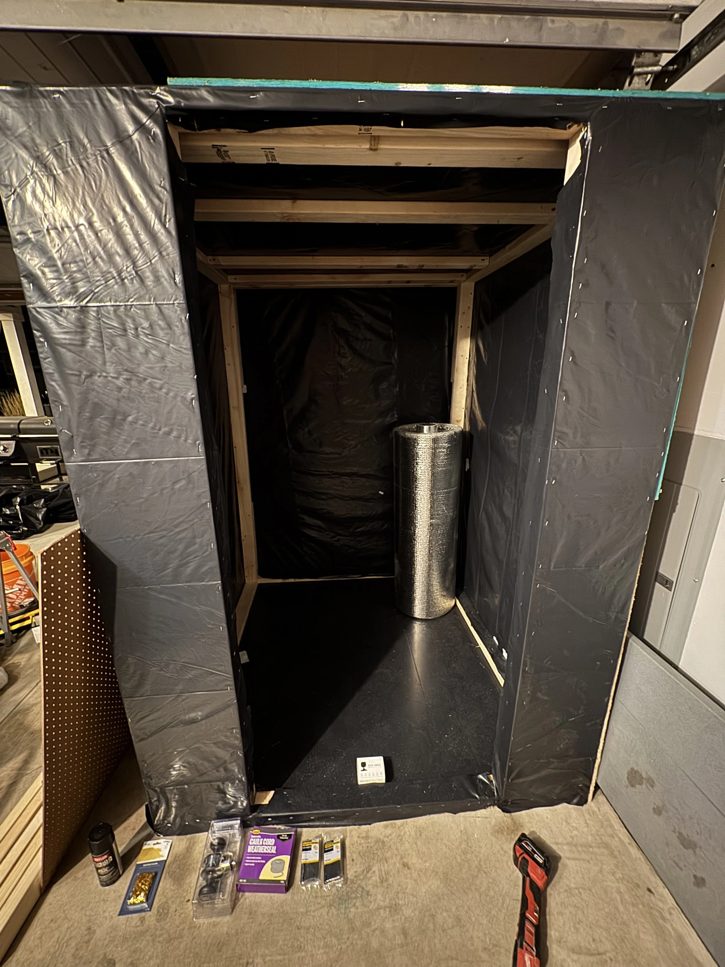 Construction of a small enclosed structure with black plastic sheathing, wooden framing, and a roll of reflective insulation inside. Construction tools and materials are on the floor, including a screwdriver, screws, and adhesive.