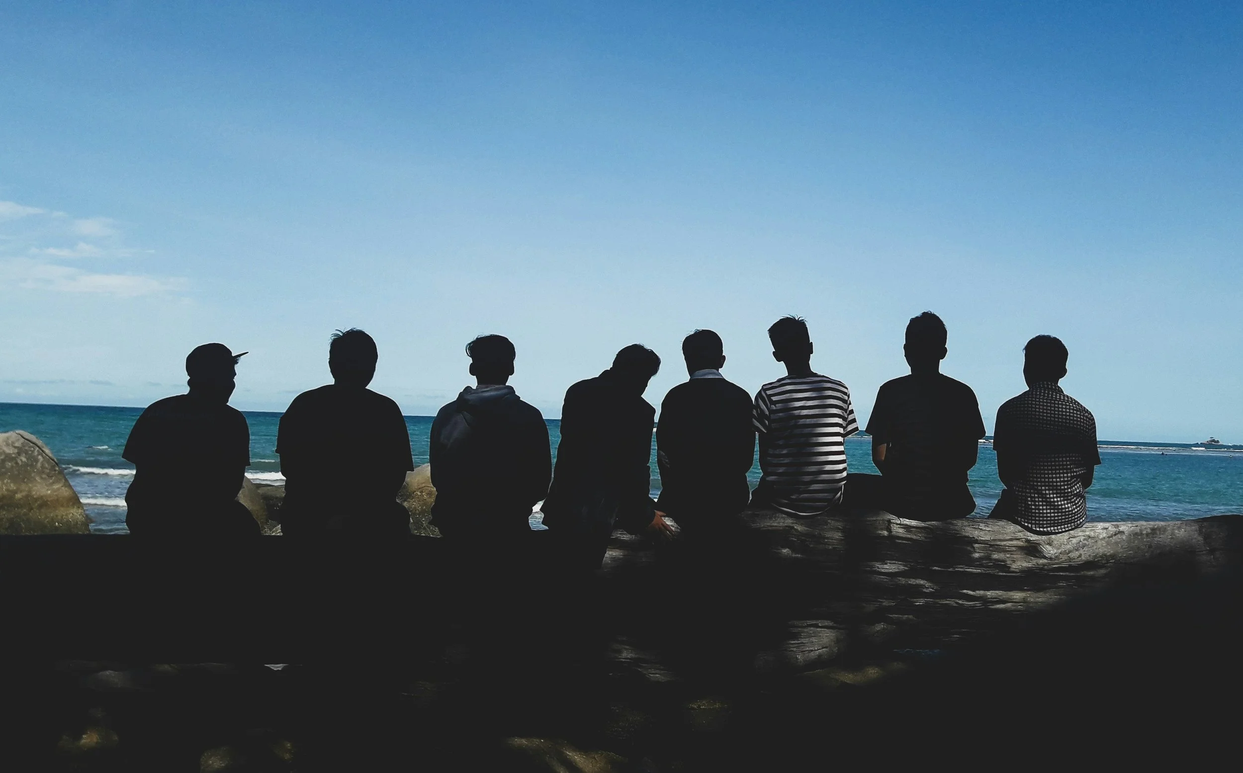 Silhouettes of eight young men sitting on a log by the beach, facing the ocean under a clear blue sky.
