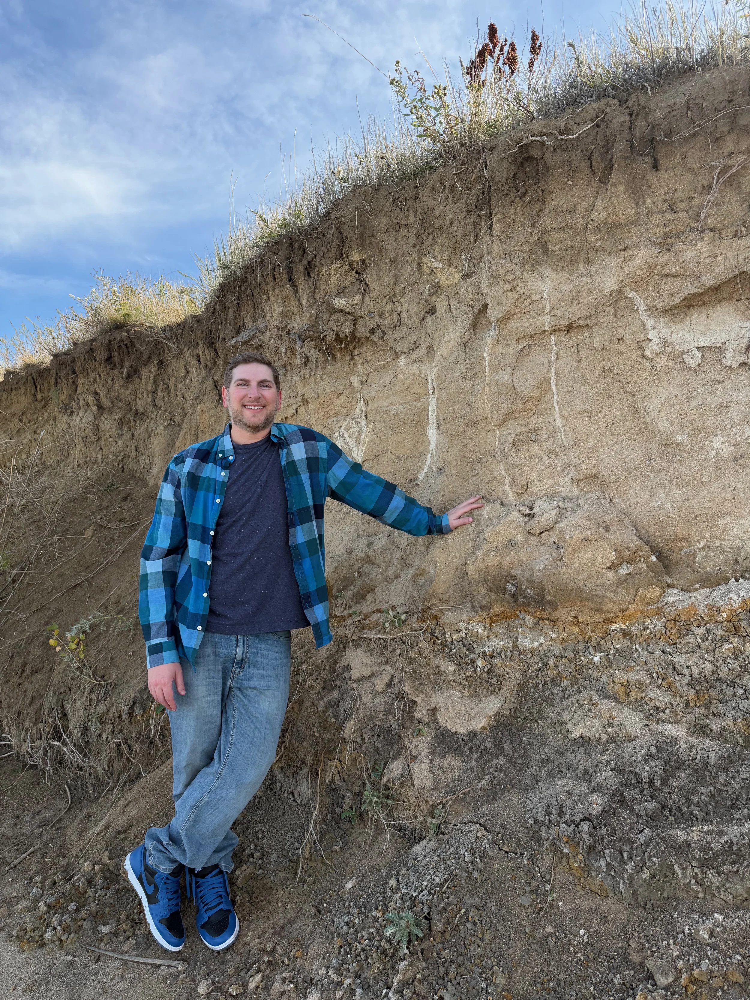 Smiling man in casual clothes touching a layered sedimentary rock formation outdoors.