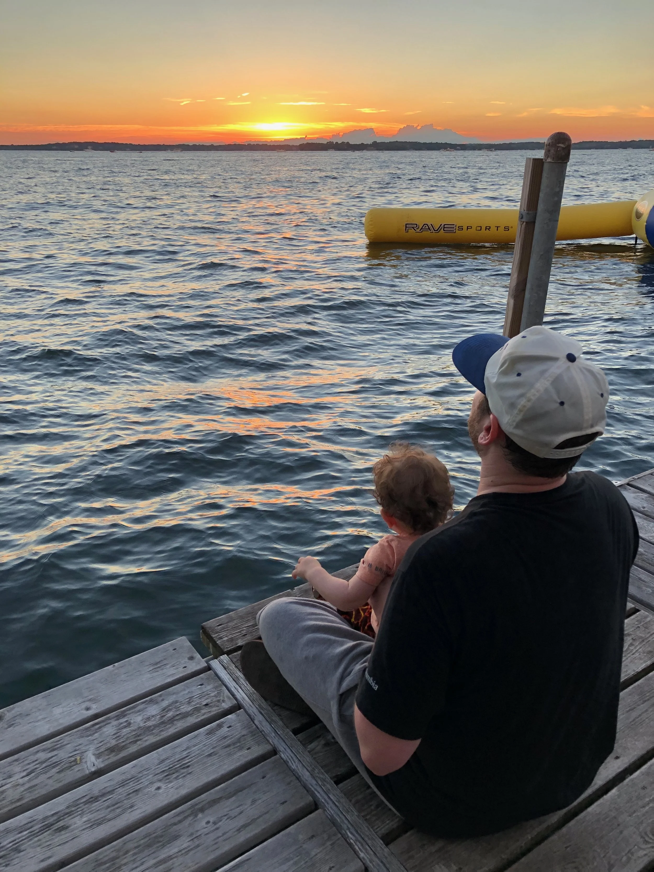 A man and a child sitting on a wooden dock by the water, watching a sunset with orange and yellow hues.