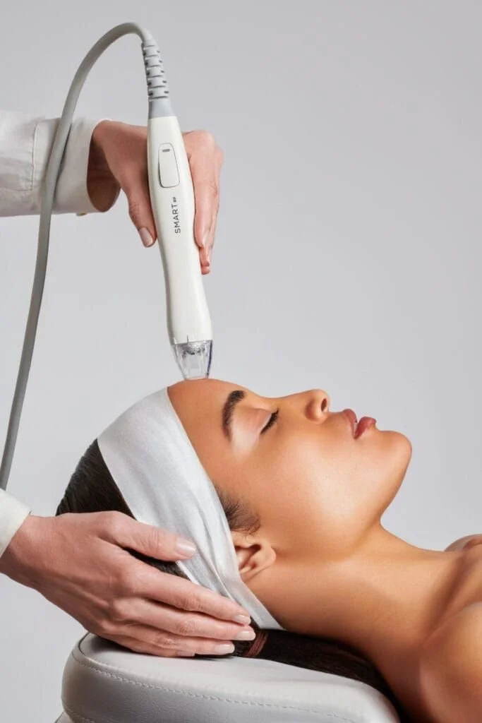 A woman receiving a facial treatment with a handheld device applied to her face, lying down with a white headband.