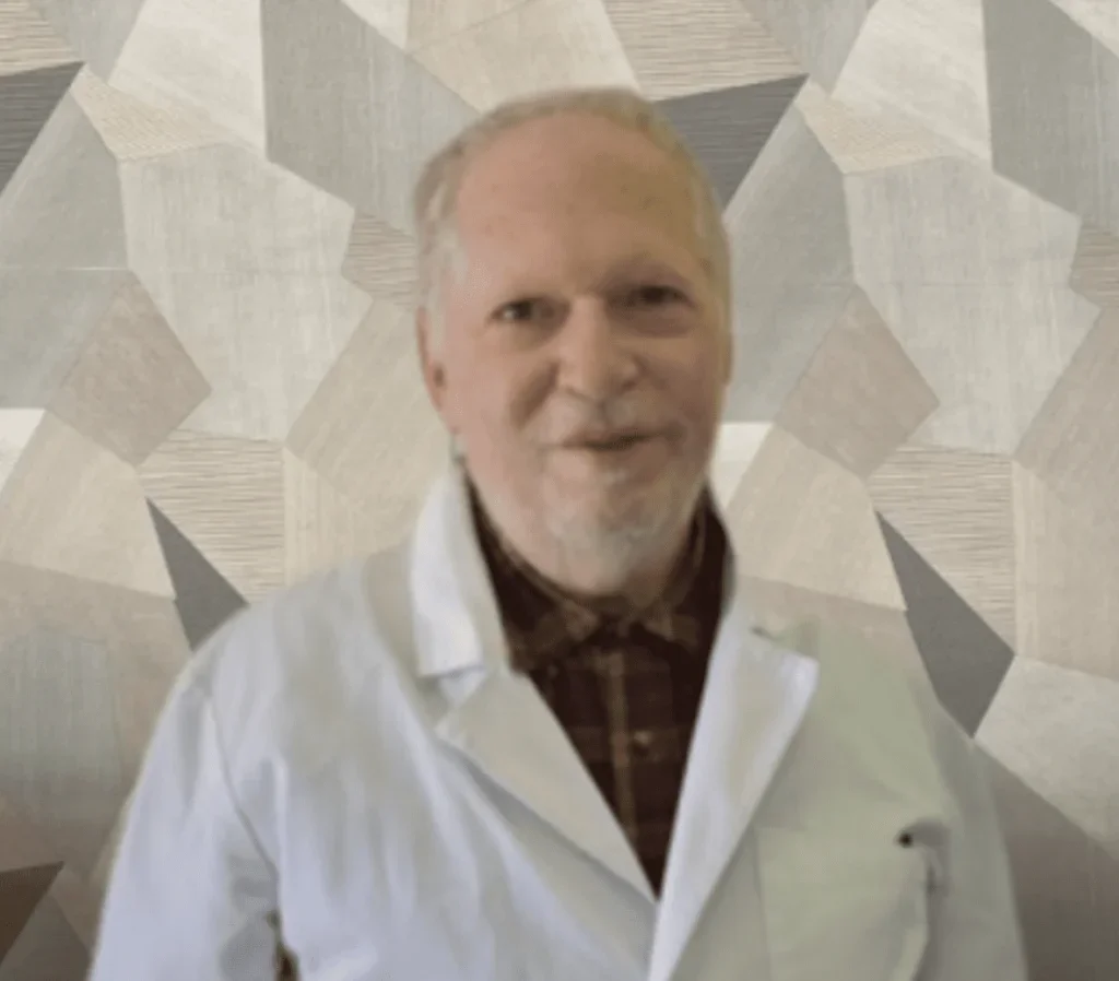 A man with gray hair and a beard wearing a white lab coat and a plaid shirt, standing in front of a patterned wall.