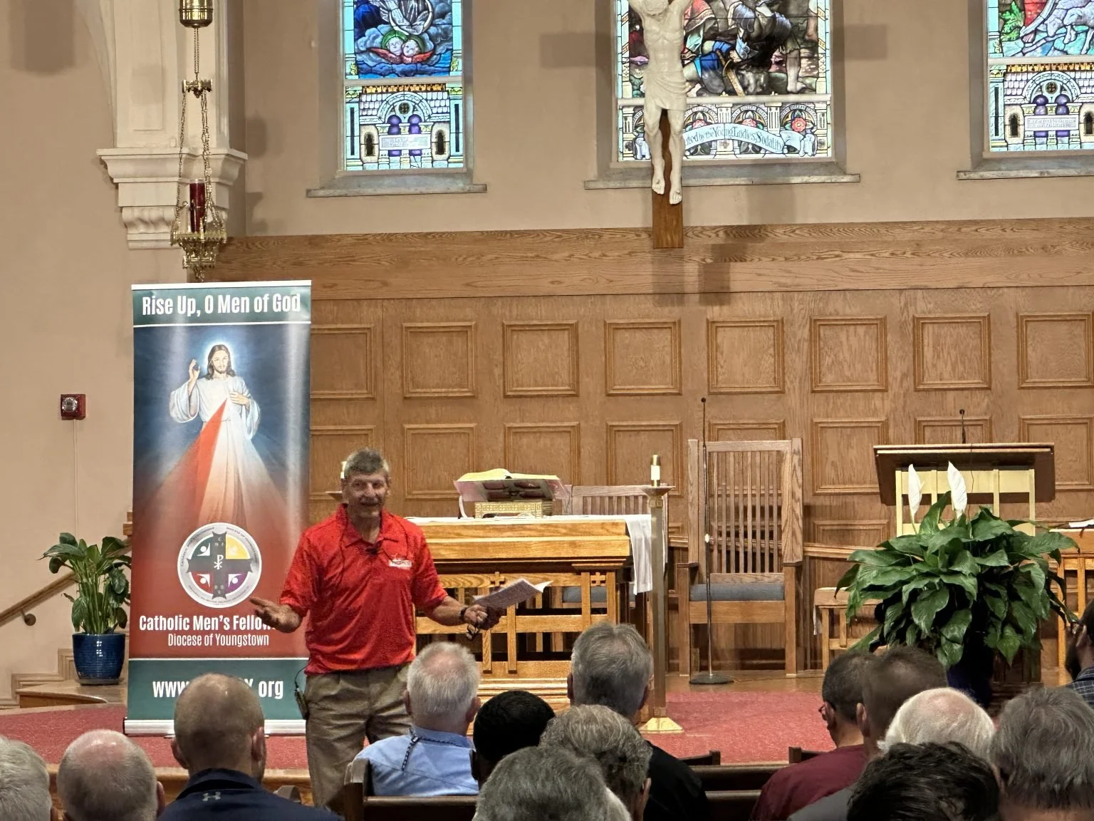 A man wearing a red shirt is speaking in front of a congregation in a church. Behind him is a banner with Jesus Christ and the words "Rise Up, O Men of God" and "Catholic Men's Fellowship, Diocese of Youngstown." The church interior features stained 