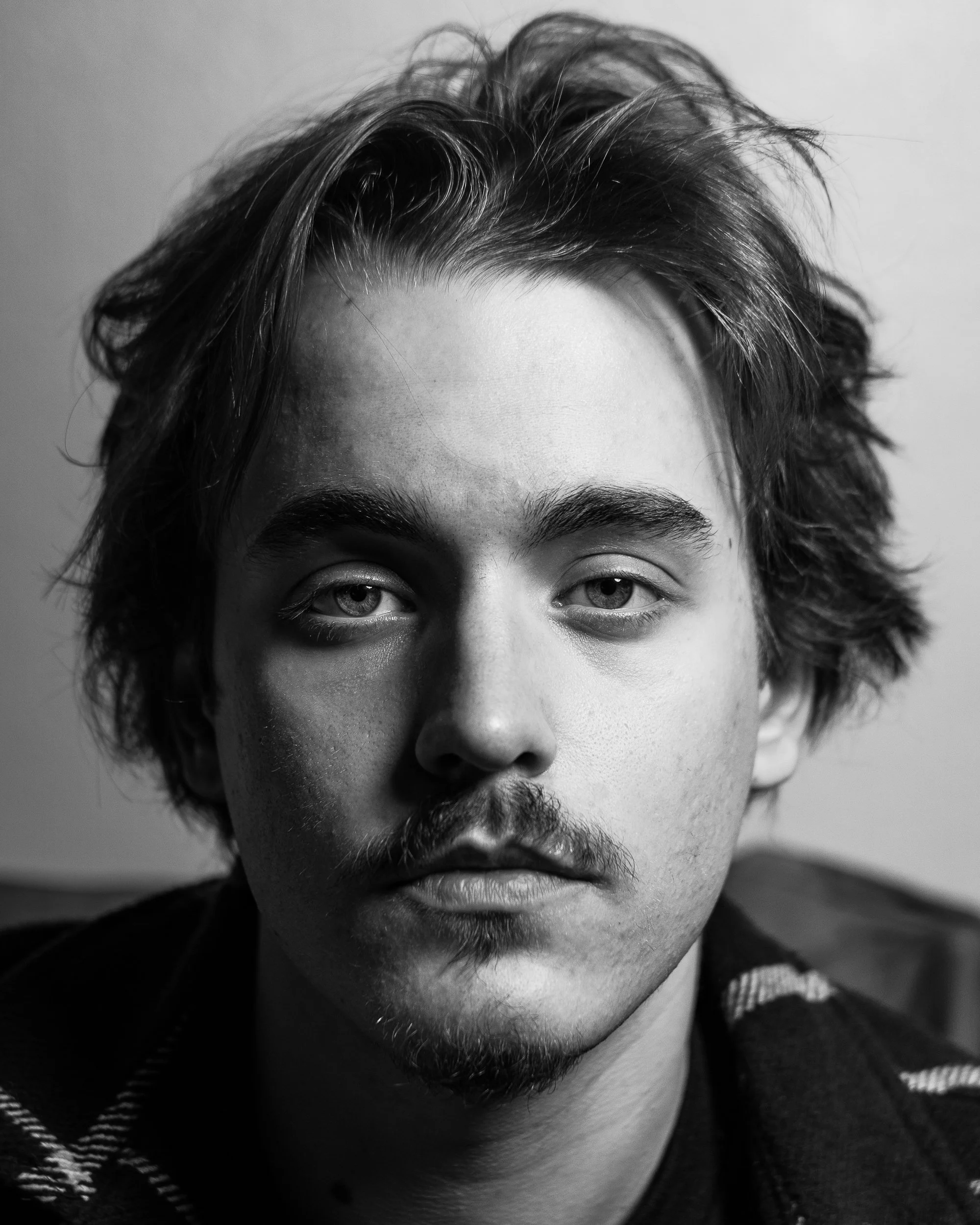 Black and white portrait of a young man with tousled hair, light eyes, and facial hair, looking directly at the camera.