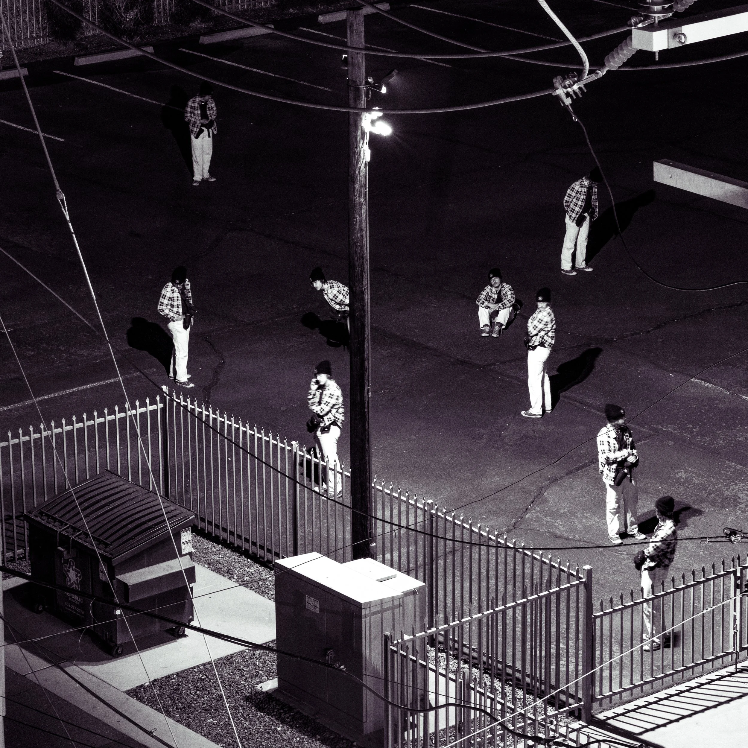 Nighttime scene on a parking lot with multiple people standing or sitting, all wearing checkered shirts, with shadows cast by overhead lights. There is a fence in the foreground and utility boxes.