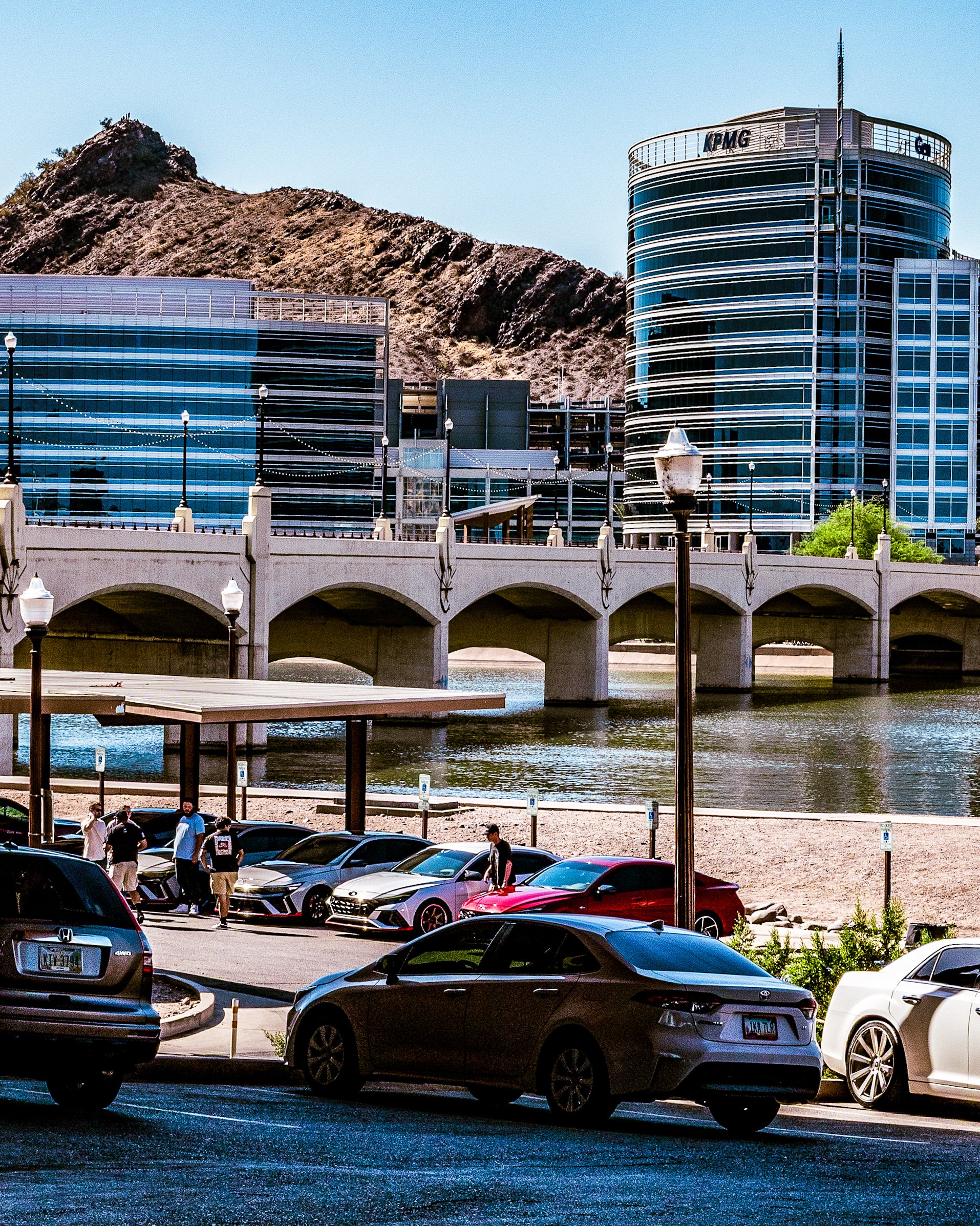 Cityscape with modern glass buildings, a bridge over water, a parking lot with cars, and a mountain in the background.