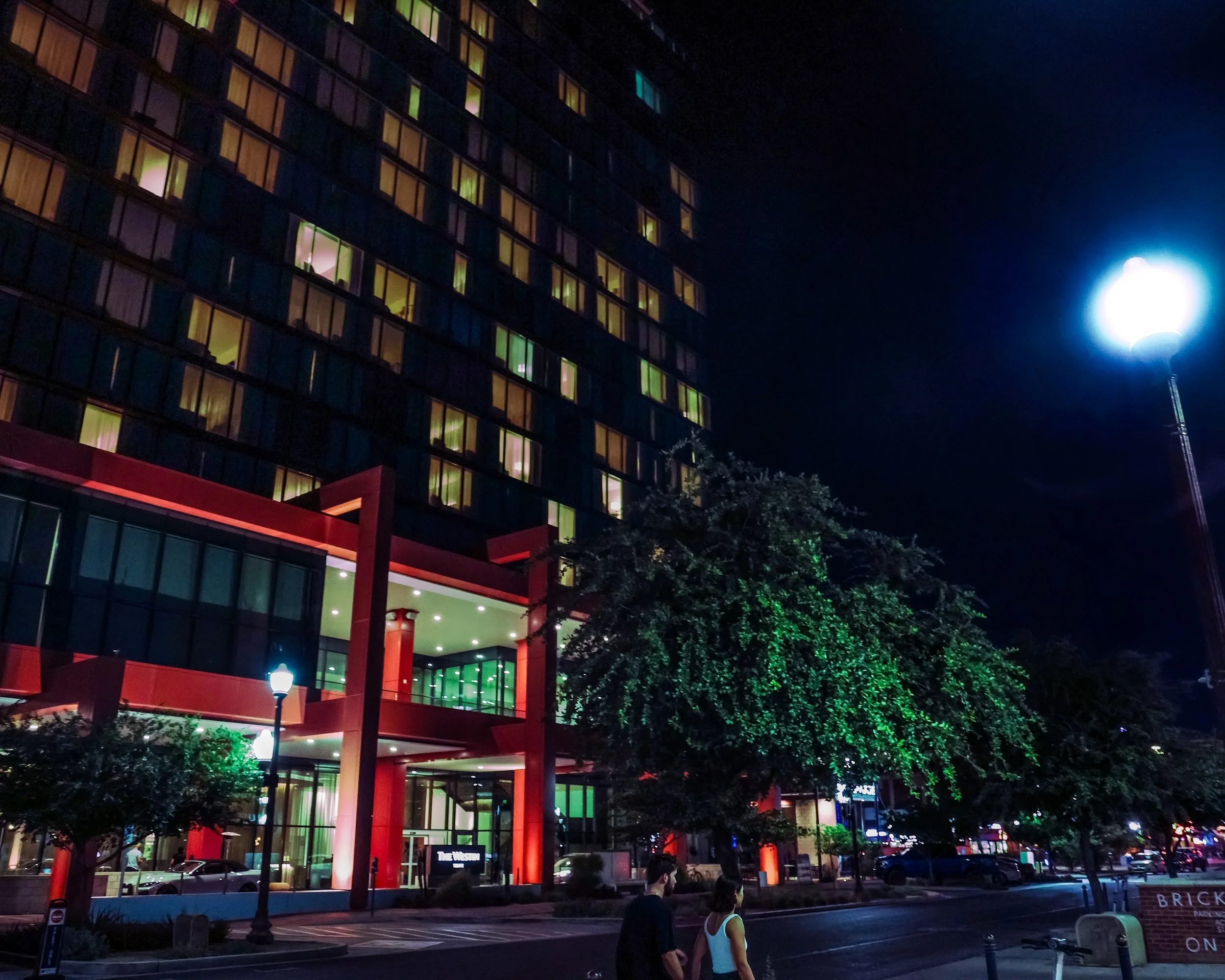 Nighttime city scene featuring a multi-story building with illuminated windows and a brightly lit entrance in red and green. There are trees, street lamps, and a few people walking on the street.