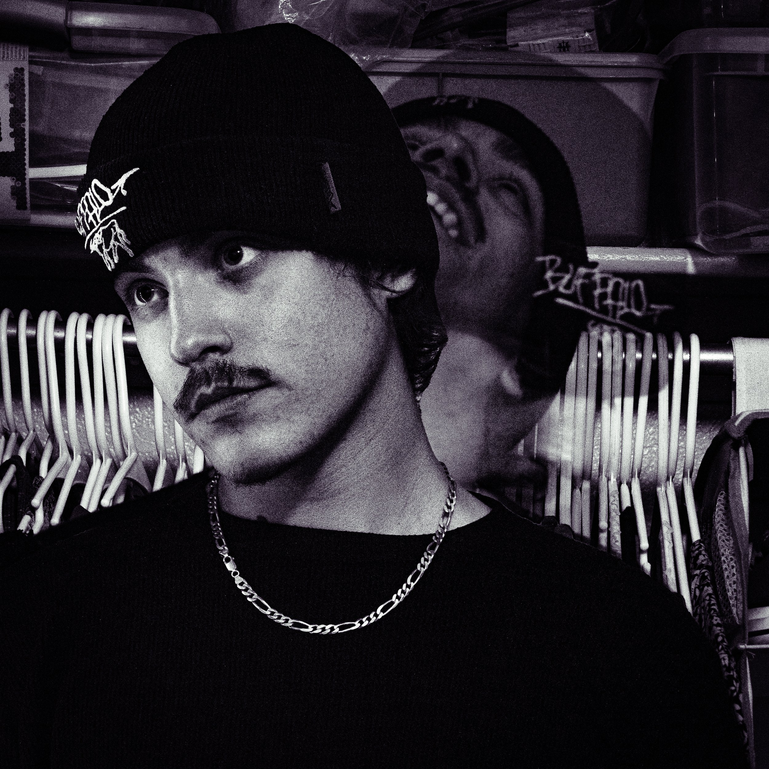 A young man with a mustache and dark hair, wearing a black beanie and black shirt, looking to the side. He has a chain necklace and is in front of a background with clothing racks and storage bins.