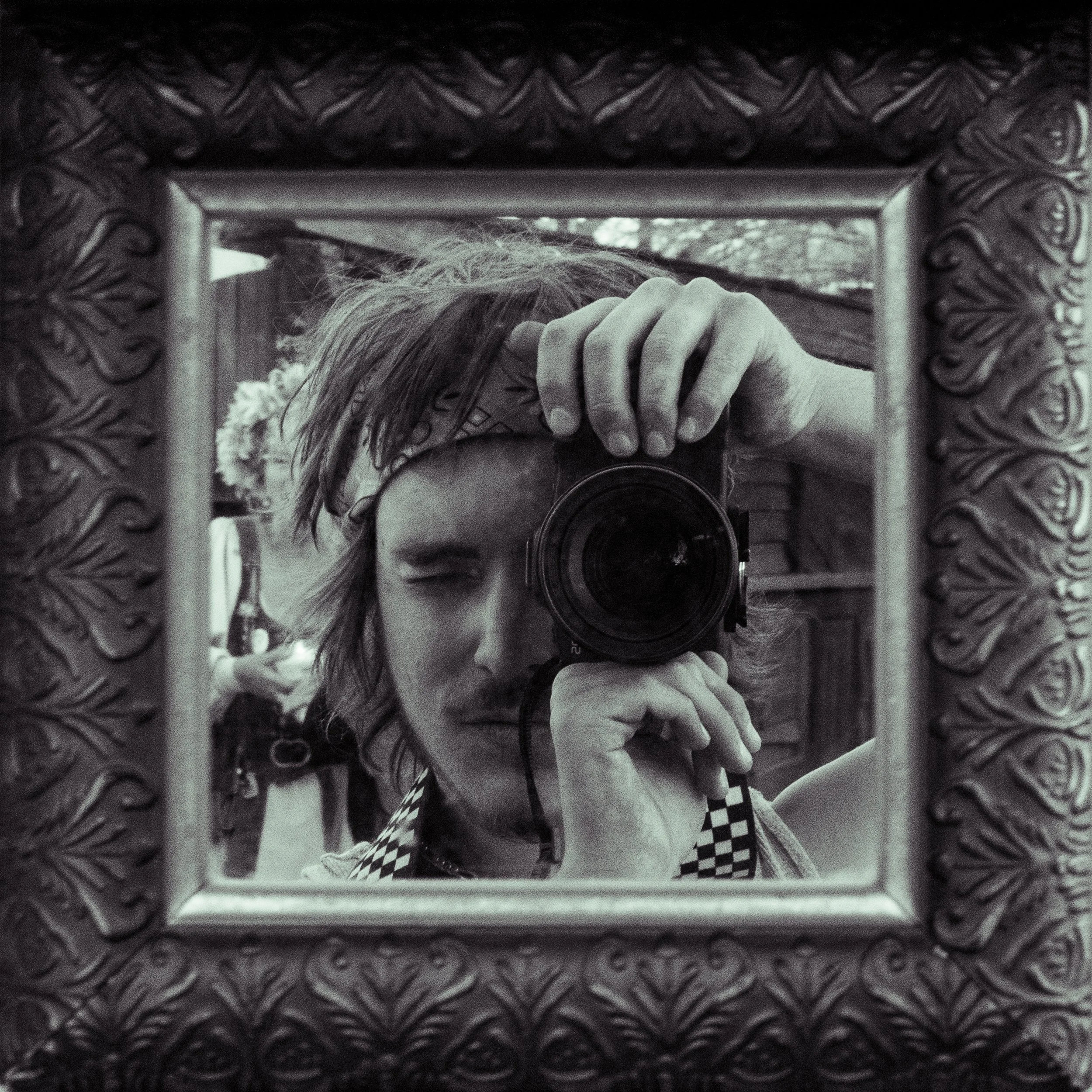 A black and white photograph of a person taking a self-portrait in a decorative mirror, with one eye closed and holding a camera up to their face, wearing a checkered shirt and a headband.