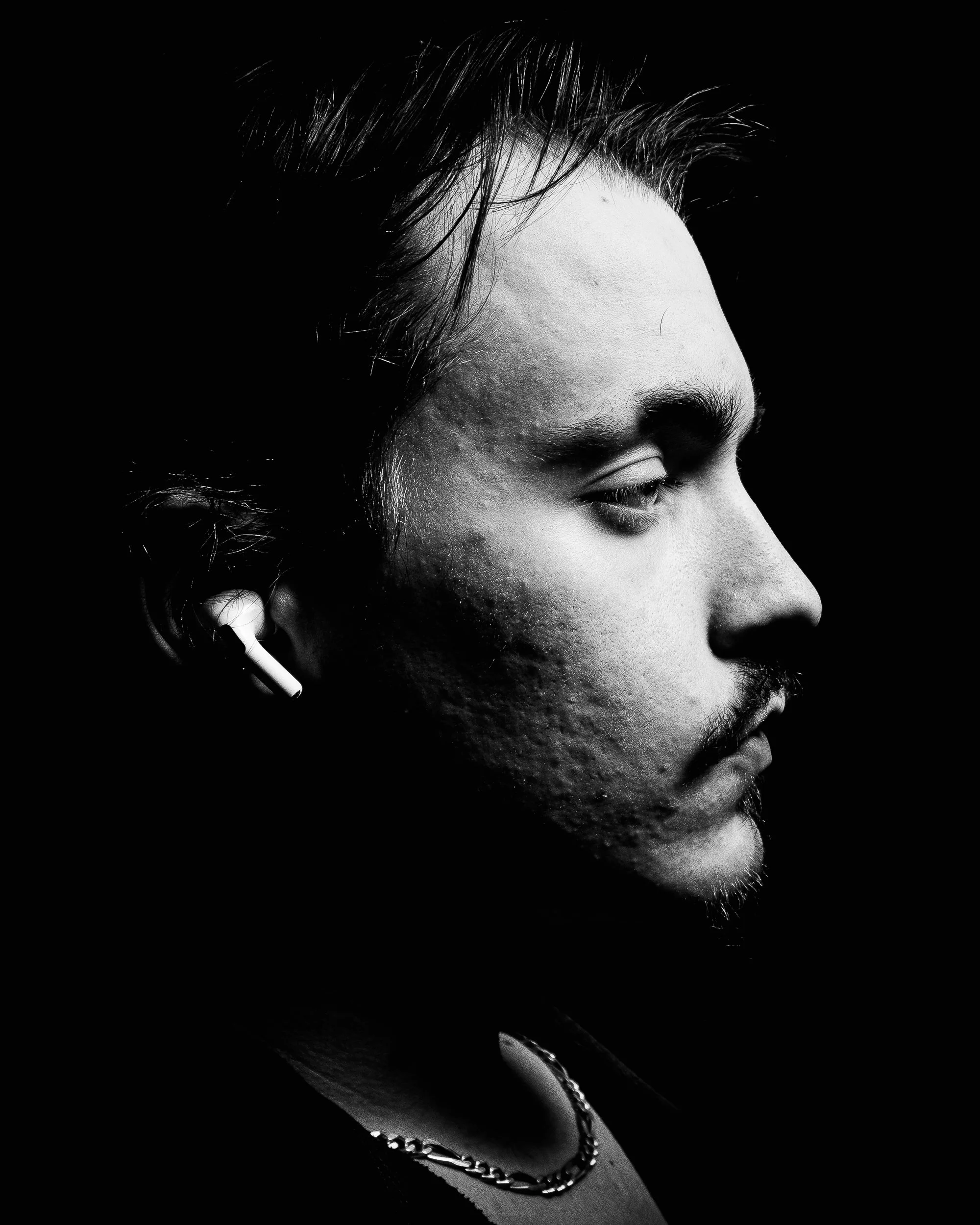 Black and white profile portrait of a man with a beard, wearing wireless earbuds and a chain necklace, against a dark background.