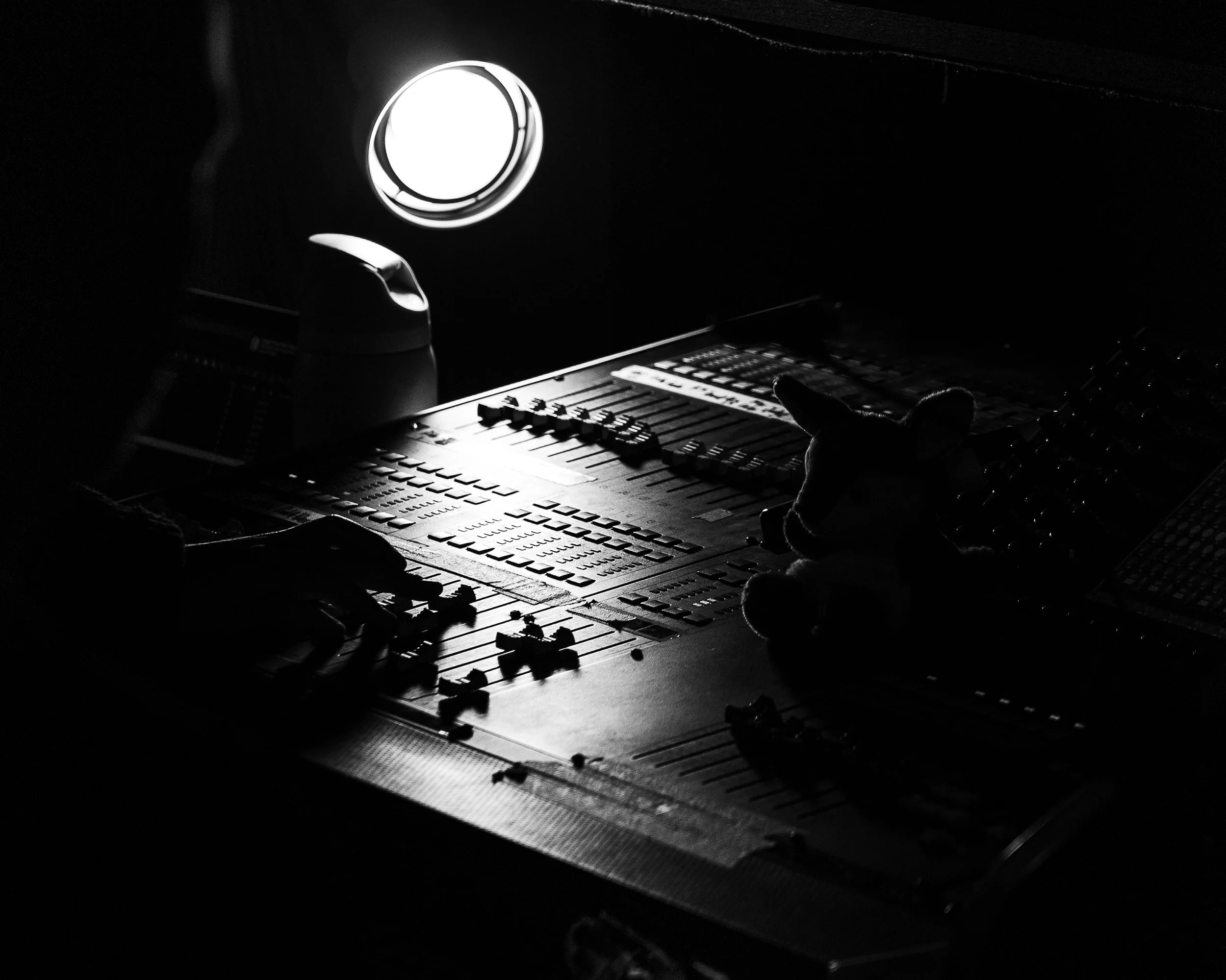 Dark photo showing a sound mixing console with a small doll on the right and a computer mouse on the left, illuminated by a bright circular light in the background.