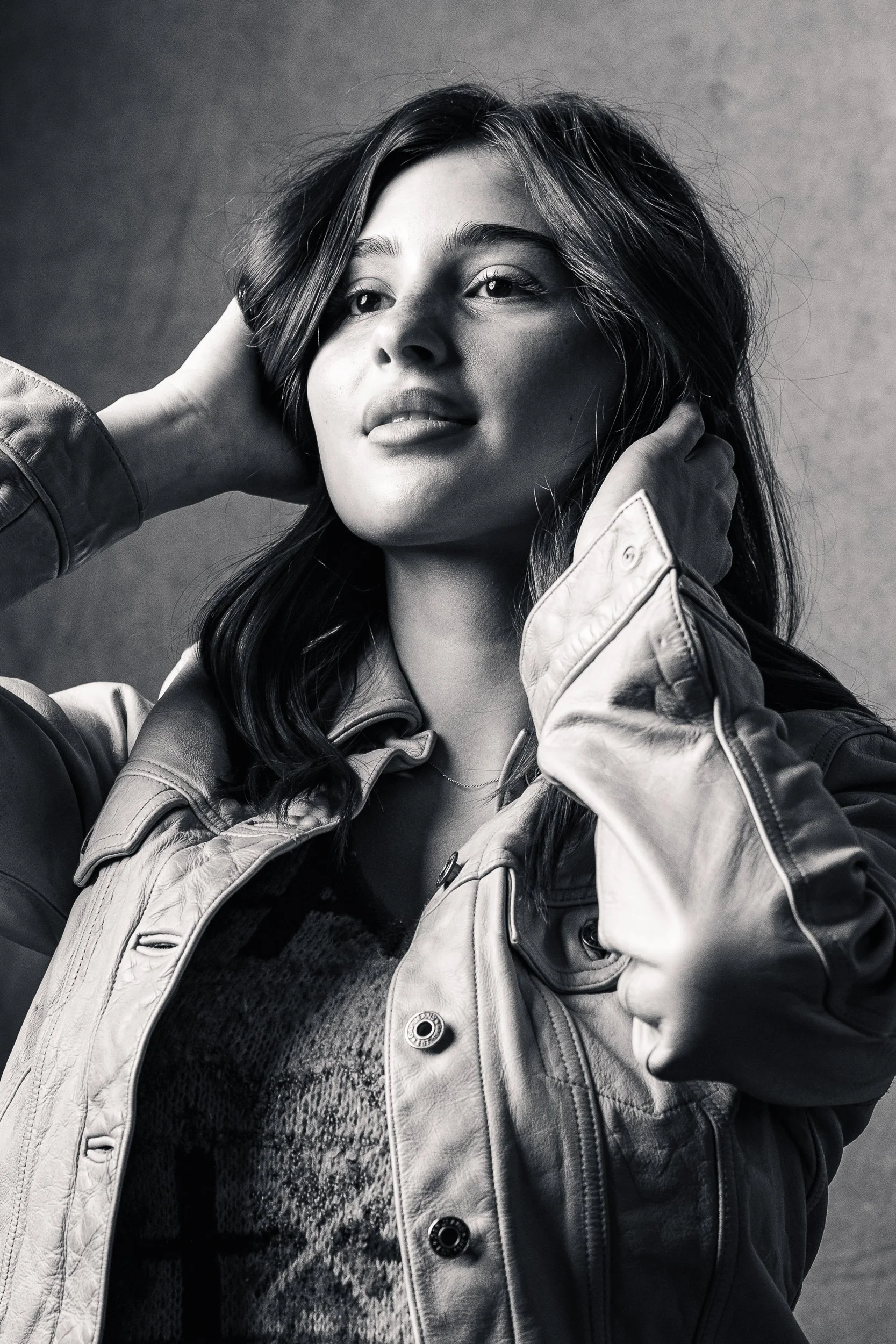 Black and white photograph of a woman with long dark hair, holding her head with both hands, wearing a leather jacket and a patterned shirt underneath.