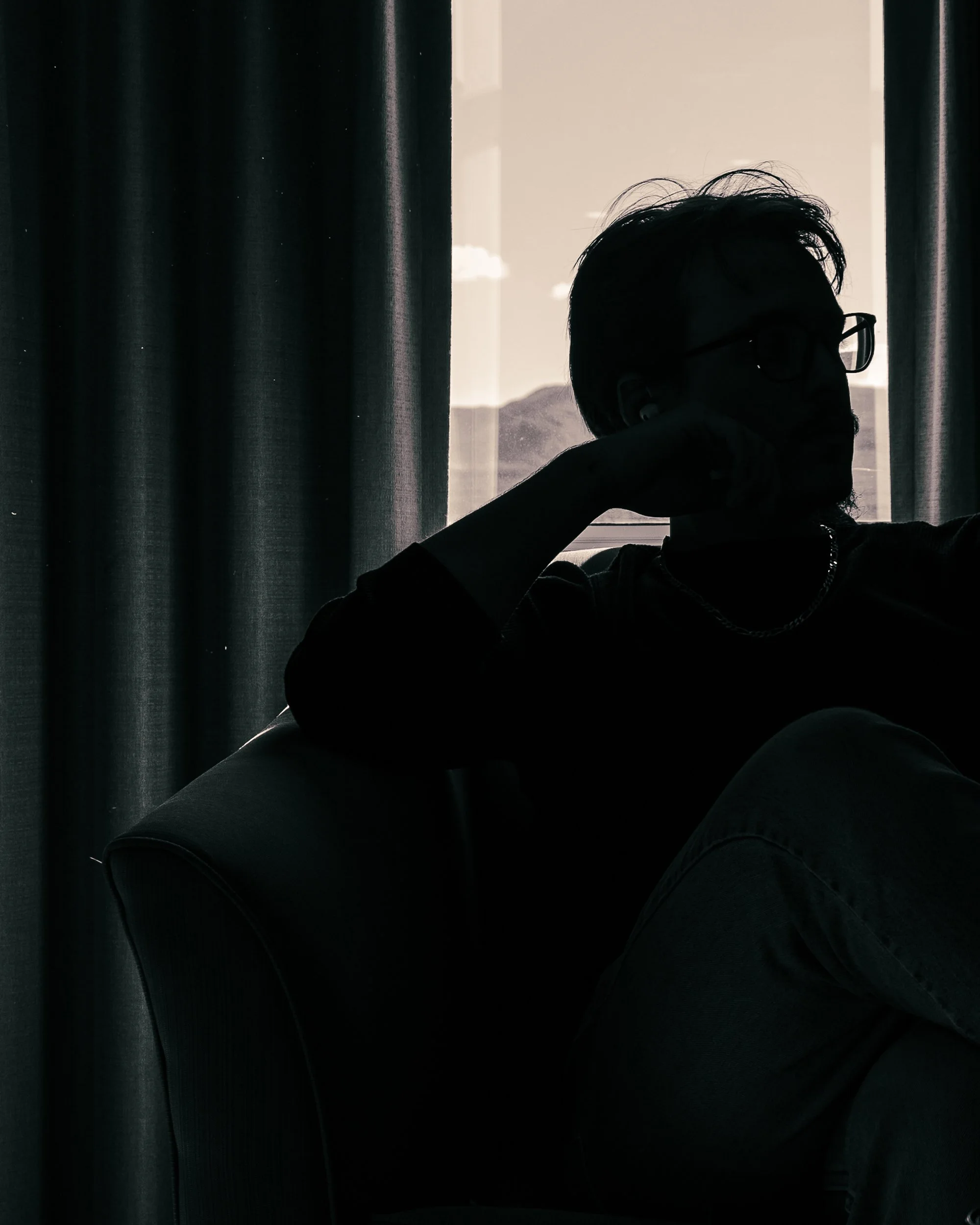 Silhouette of a man with glasses and a chain necklace sitting on a chair near a window with curtains, backlit by natural light.
