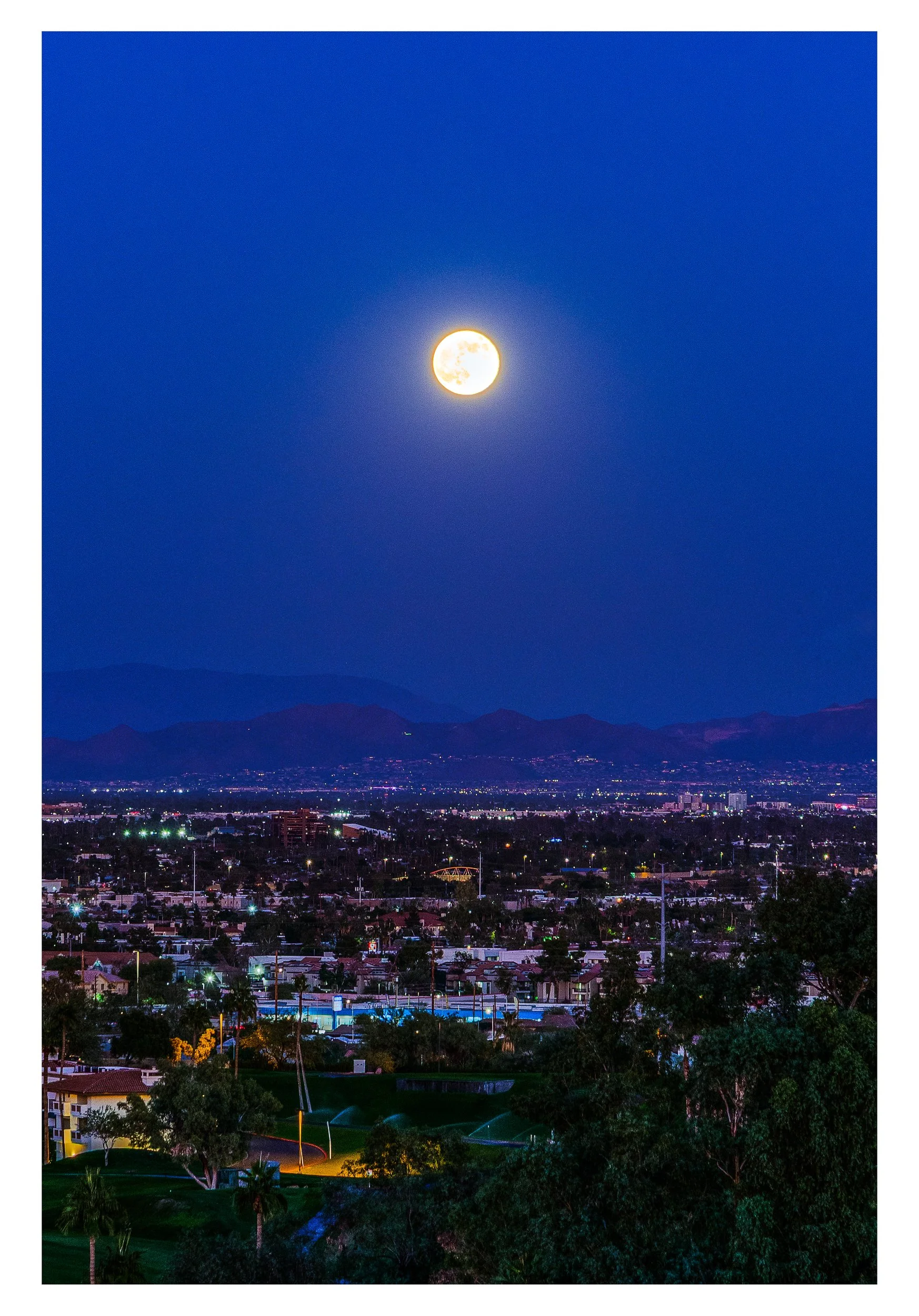 Nighttime cityscape with a full moon in a clear blue sky, mountains in the background, and illuminated buildings and streets in the foreground.