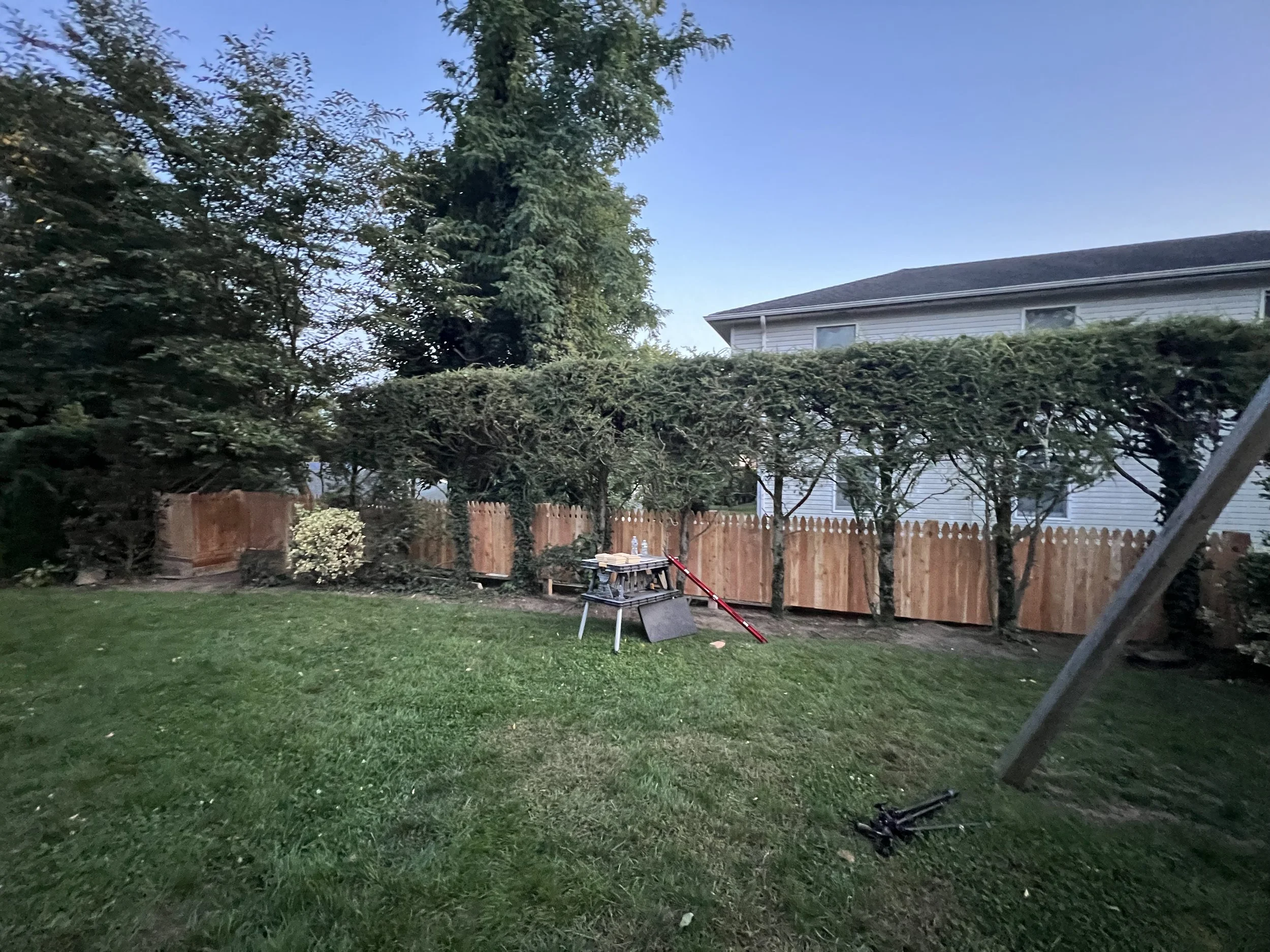 A backyard scene at dusk with a green lawn, a wooden fence, and trees. A picnic table holds tools, and a red spirit level leans on the table. A avoidable pole is toppled on the ground.