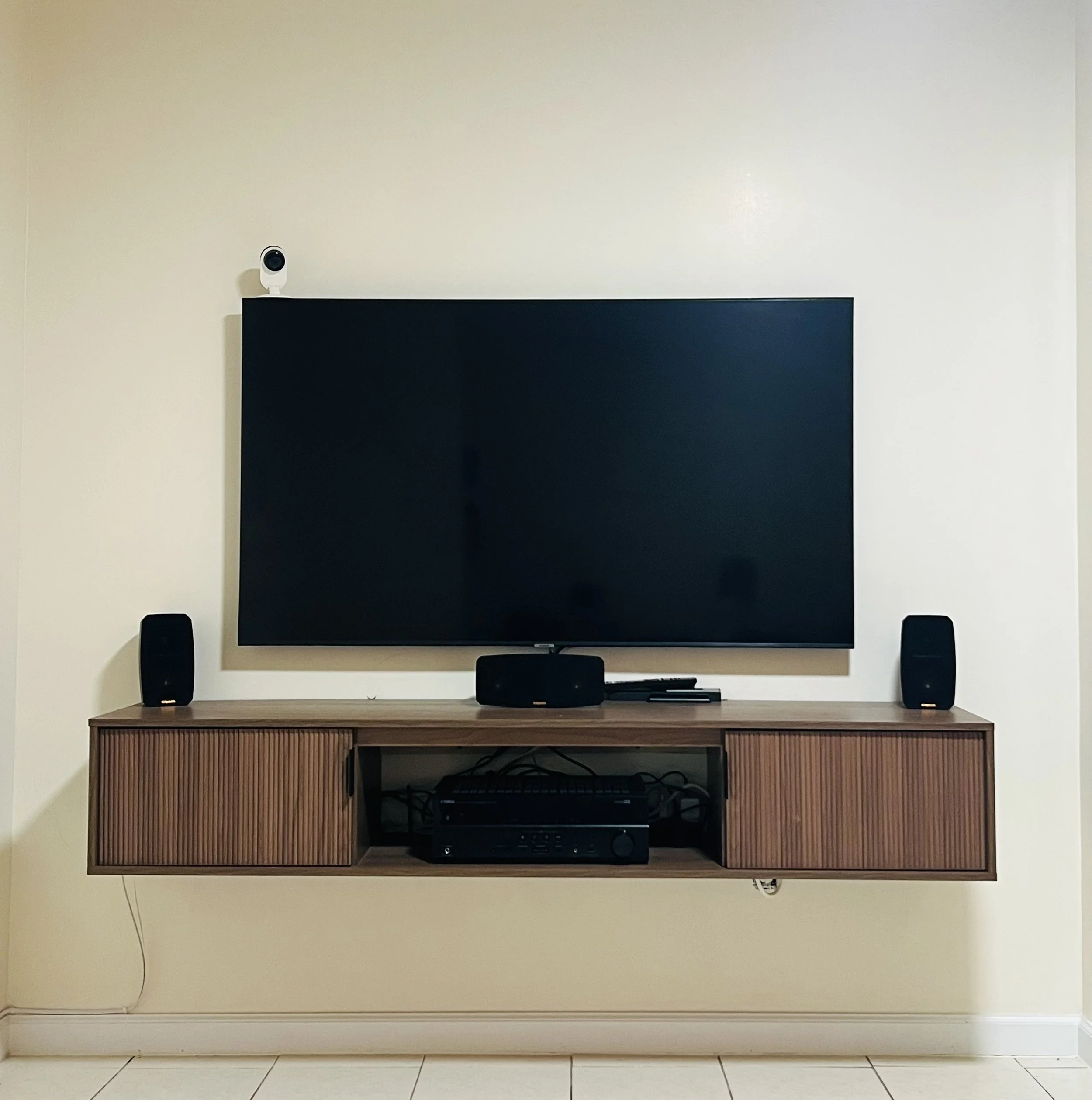 Living room entertainment setup with a flat-screen TV mounted on a wall, two black speakers on either side, a soundbar and remote on a wooden wall-mounted console, with an AV receiver beneath the console.