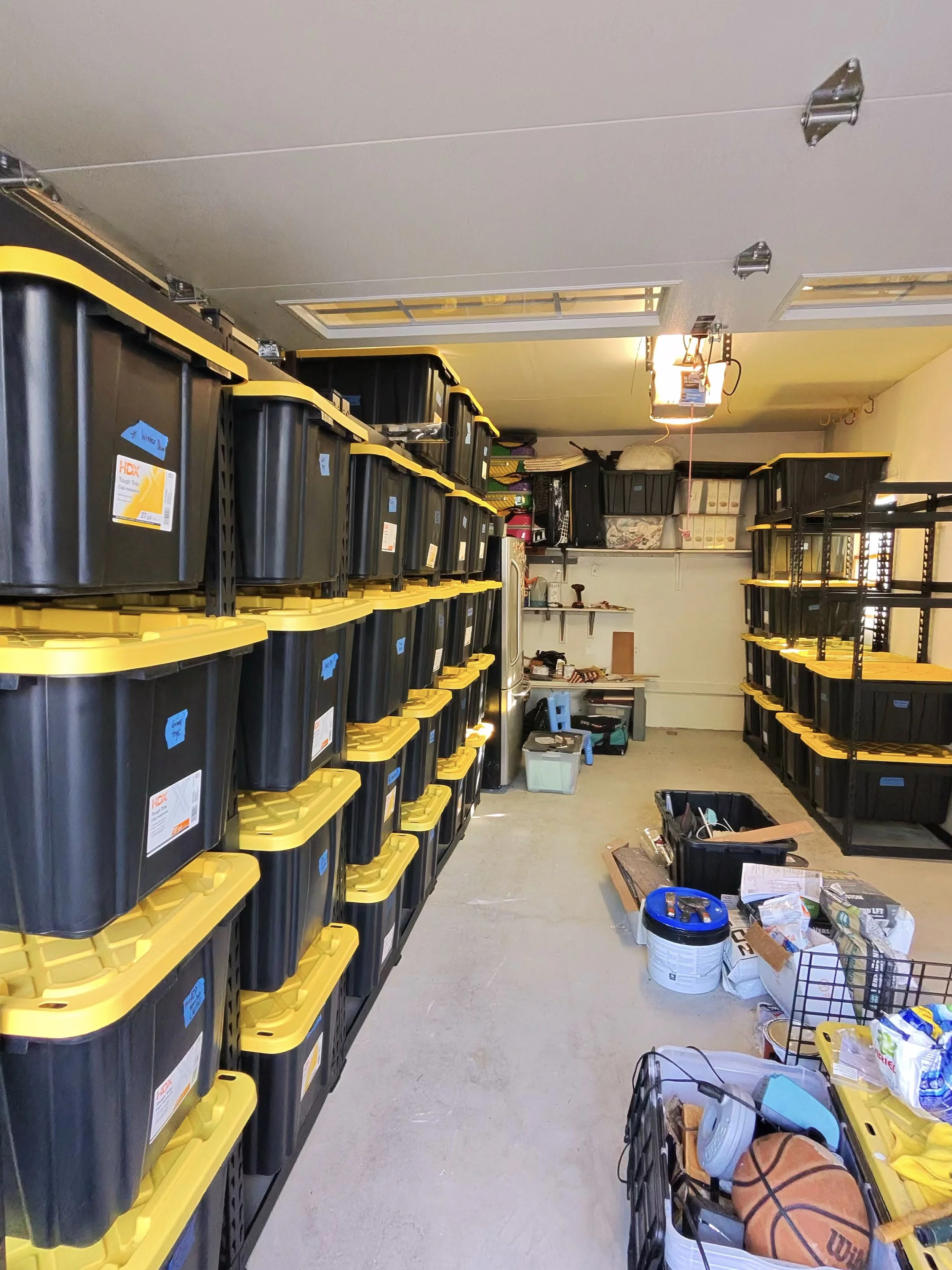 Garage with organized black storage bins with yellow lids on metal shelves, various tools and items scattered on the floor, and a closed garage door