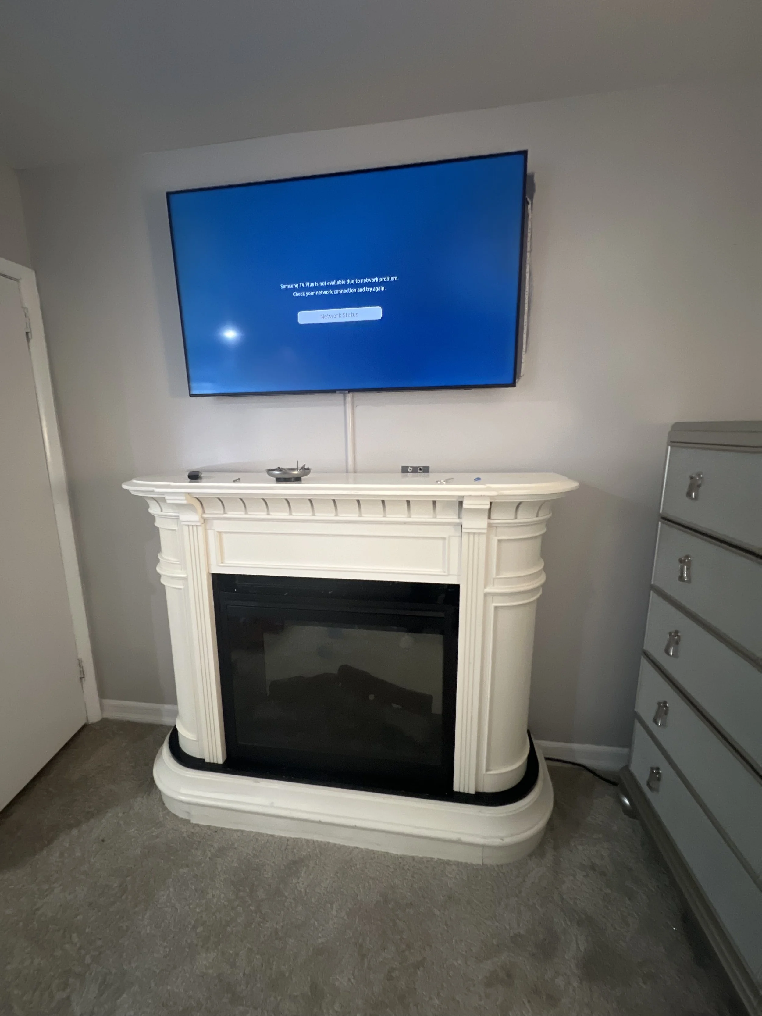 A flat-screen TV mounted on a white wall, displaying a network error message. The TV is placed above a decorative white fireplace mantel, which houses an electric fireplace insert. To the right of the mantel, there is a silver dresser with multiple drawers.