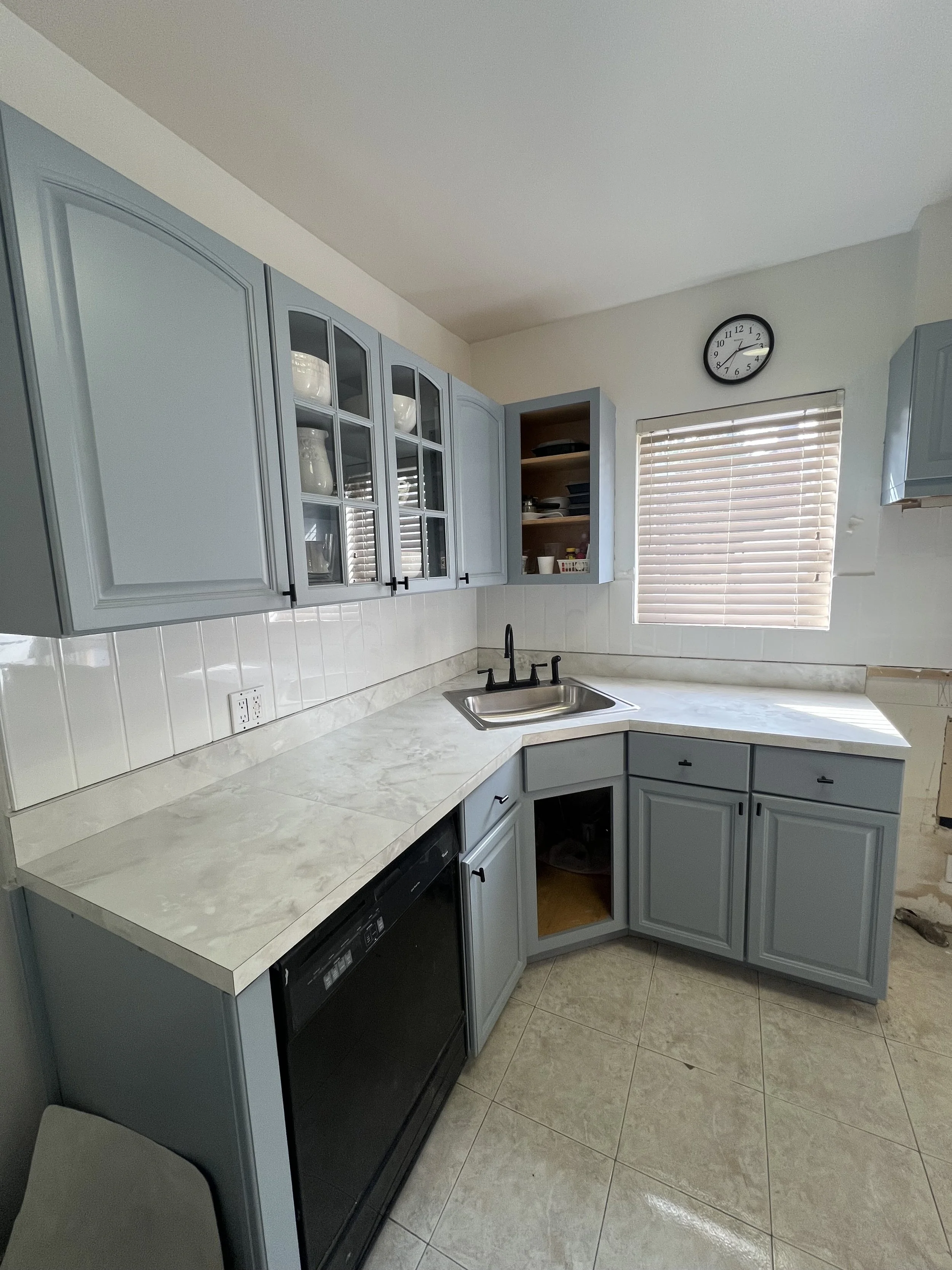 Kitchen with light blue cabinets, marble countertop, white tiled backsplash, black faucet, small sink, partial view of dishwasher, window with blinds, wall clock, open shelf with dishes and supplies.