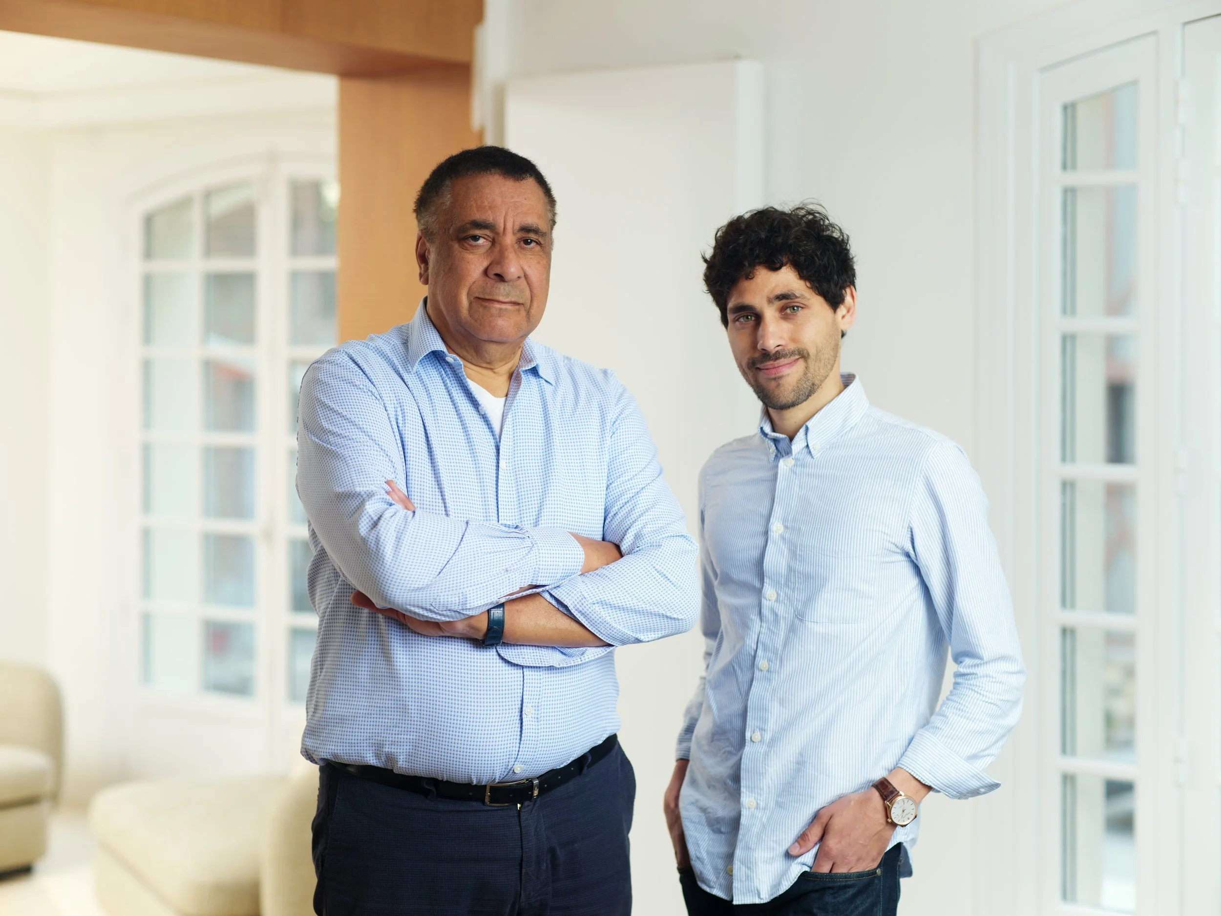 Two men in business casual attire standing inside a bright room with large windows, one with arms crossed and the other with hands in pockets.