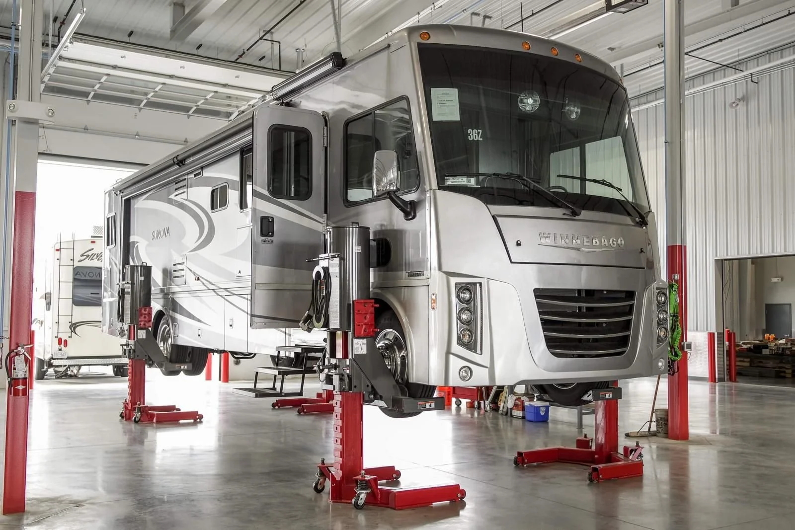 Inside a large workshop, a Winnebago RV is elevated on red hydraulic lifts, with tools and equipment around it.