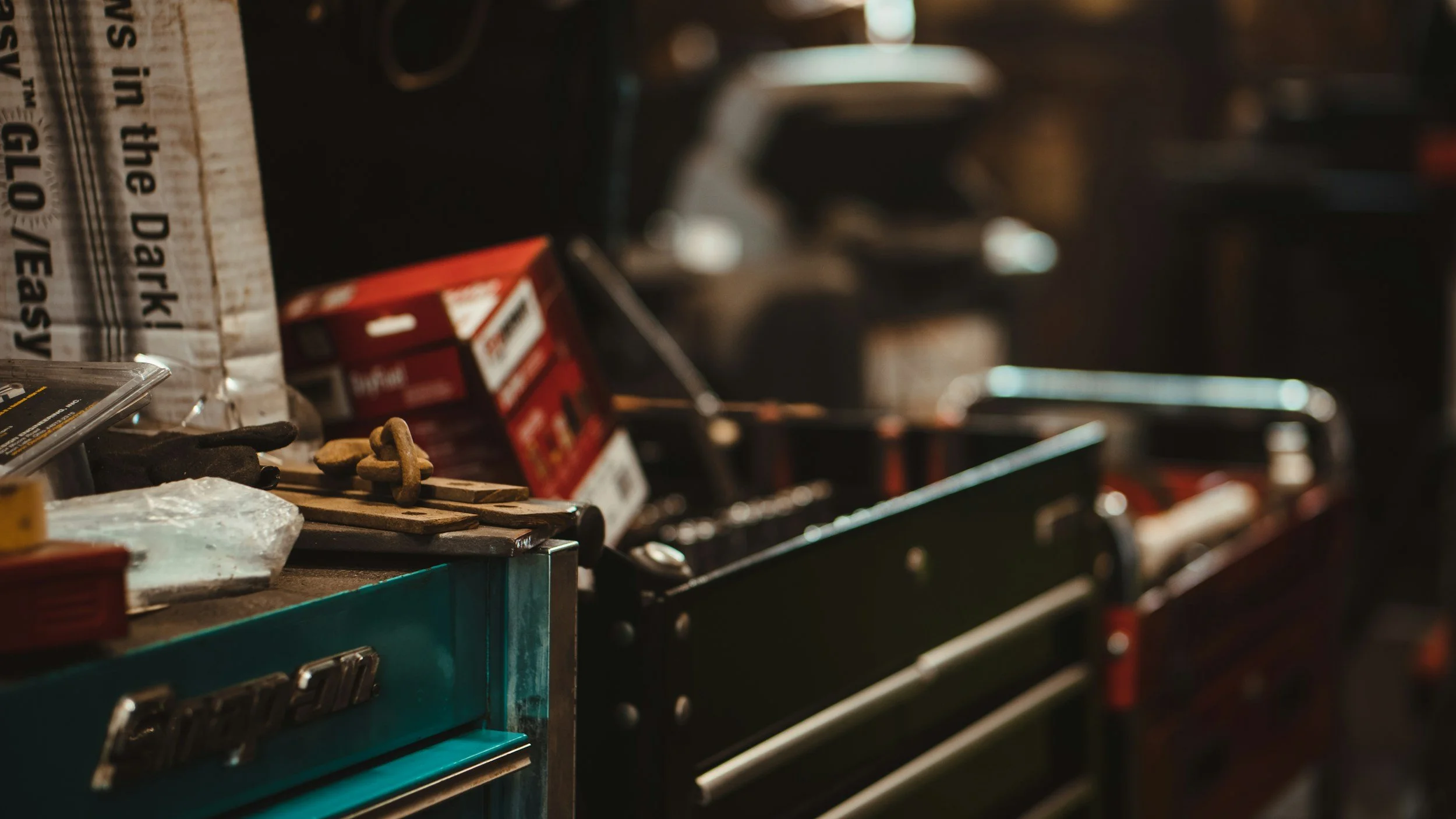 Close-up of a cluttered tool chest with various tools and small items on top, including a coiled wire, red and black boxes, and small metallic parts, in a workshop setting.
