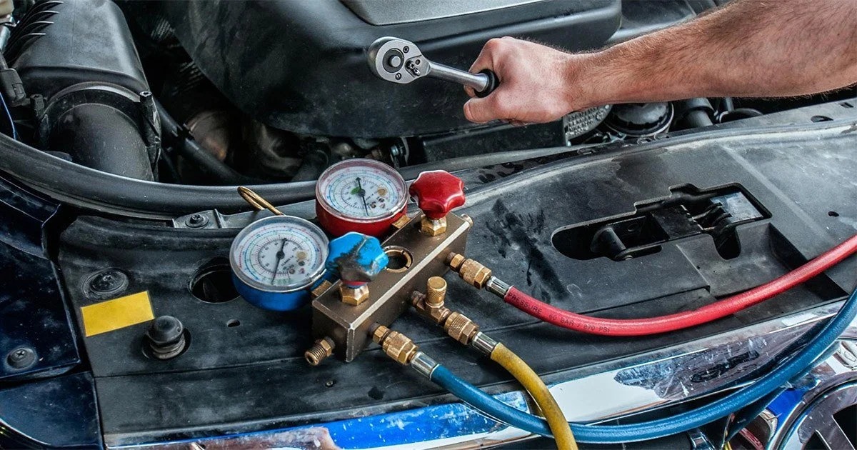 Close-up of an automotive air conditioning service port with a mechanic's hand using a tool to connect or disconnect hoses, gauges, and a manifold gauge set on a car's engine bay.
